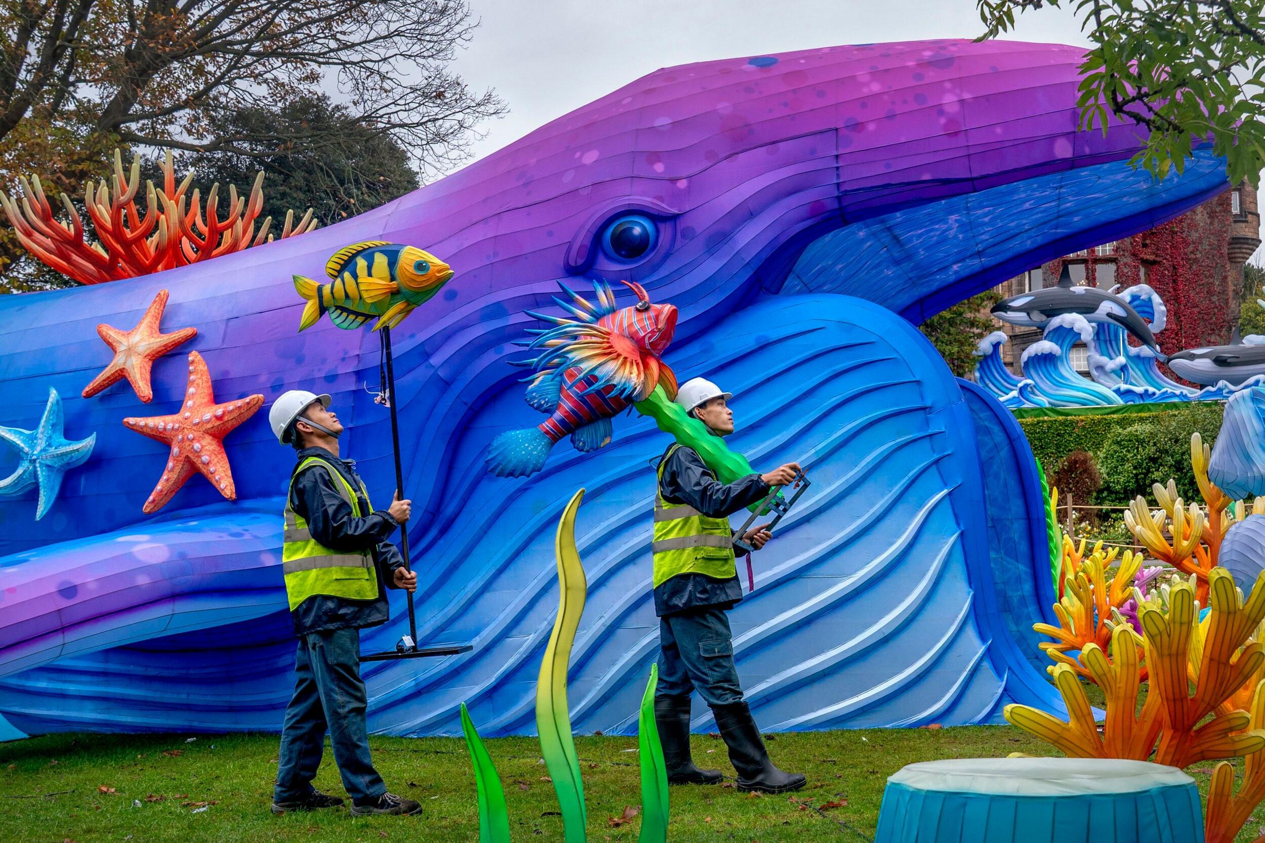 Two men in hard hats and fluorescent jackets carry lanterns shaped like fish above their heads. Behind them there is a large model of a whale with starfish and coral-shaped lanterns