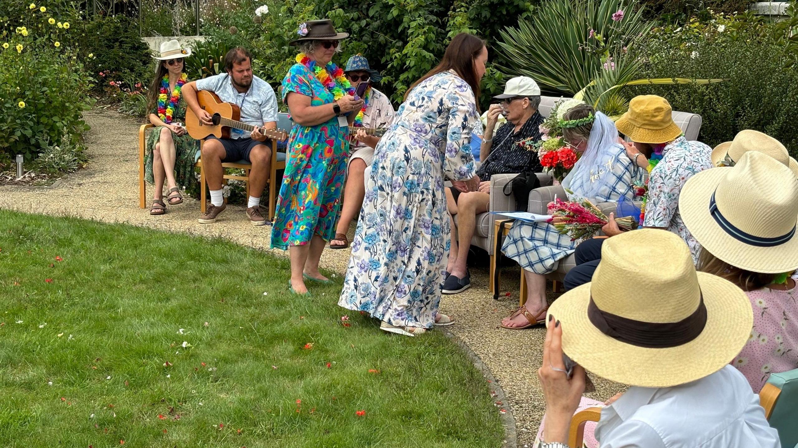 People sit in a semi-circle of chairs in the garden as Alison signs the wedding document with David sat next to her. Her son has his hand on her shoulder to support her. A man plays an acoustic at the edge of the circle 