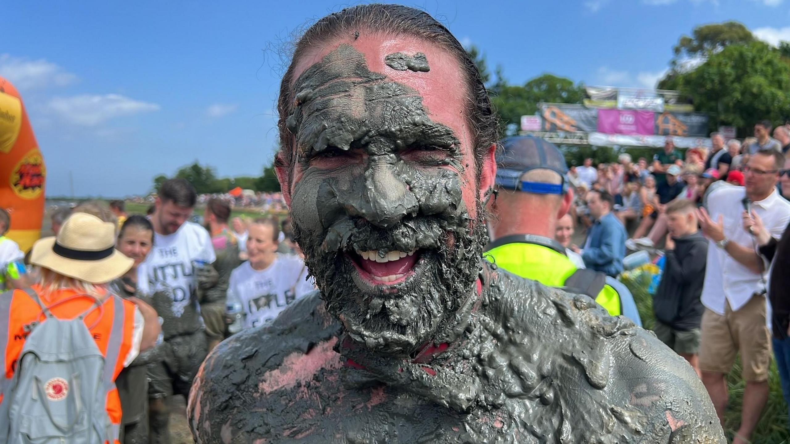 A laughing, bearded man with brown hair covered in mud with crowds behind him.
