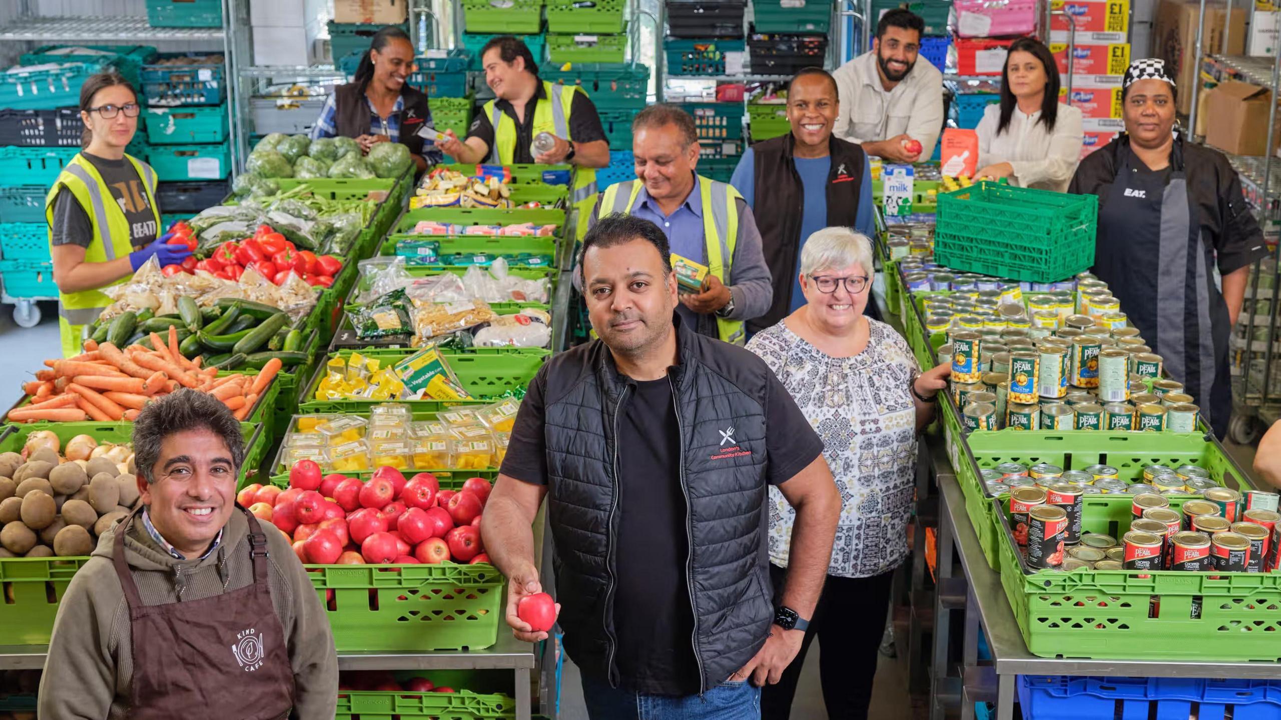 Taz Khan, a man with dark hair wearing a black gilet and t-shirt, stands in front of green trays of colourful produce and tinned food with his team in a warehouse