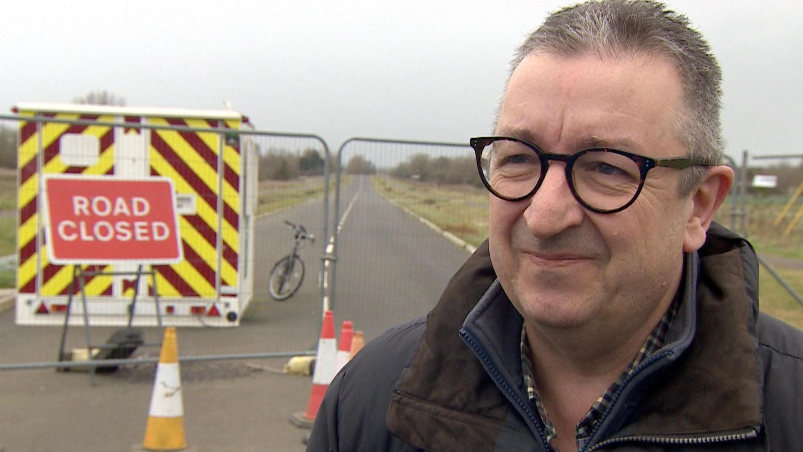 Gary Sumner is wearing black glasses and has short grey hair. He is standing beside a red and white sign that says 'road closed'. There is an empty road behind him, with a metal fence across it. There are several traffic cones and a temporary structure on the road with large yellow and red stripes.