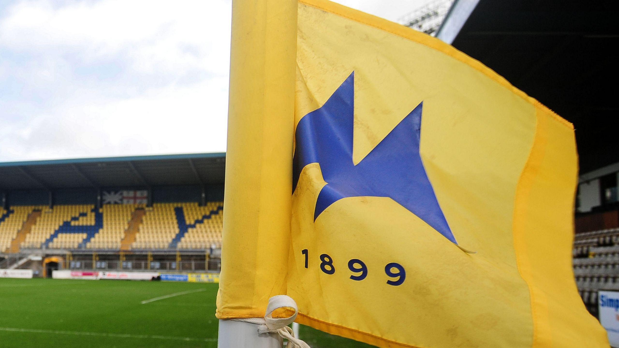 Torquay United's Plainmoor stadium. A yellow and blue flag with "1899" is in the foreground and the pitch and stands in the background.