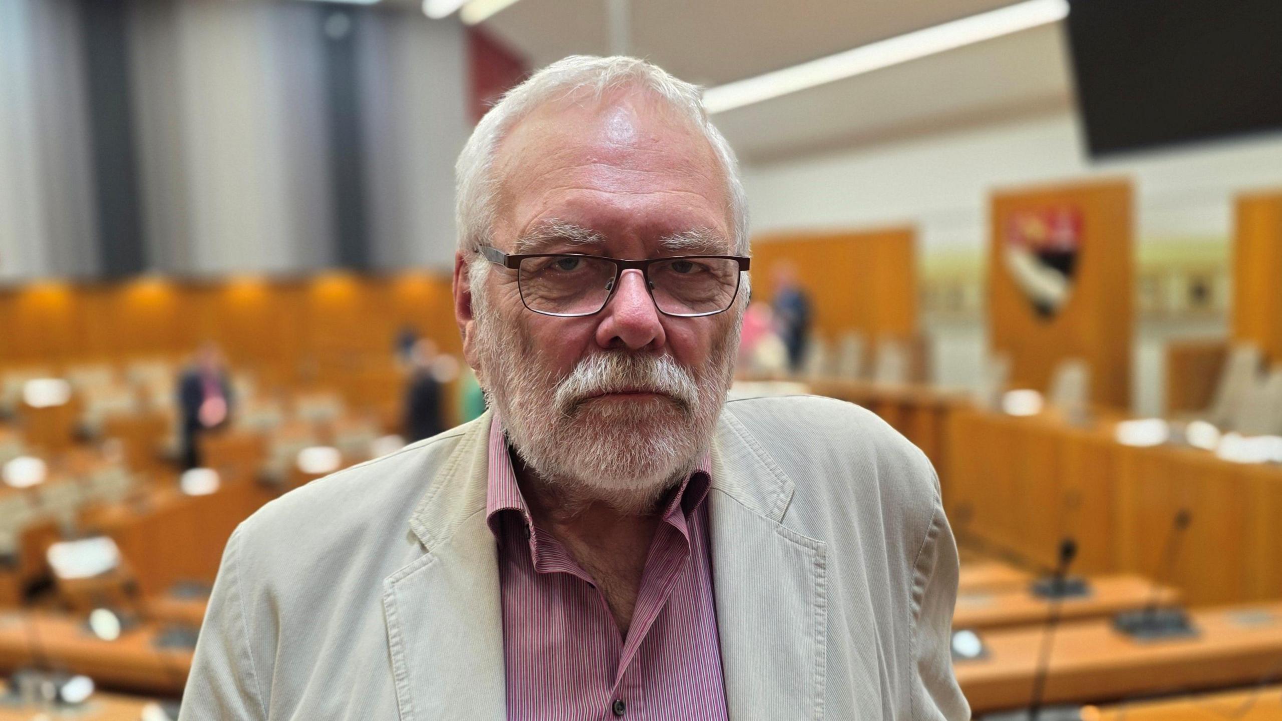 Steve Morphew is standing in a council chamber. He's wearing a light coloured linen jacket over a red shirt, with white patterning.