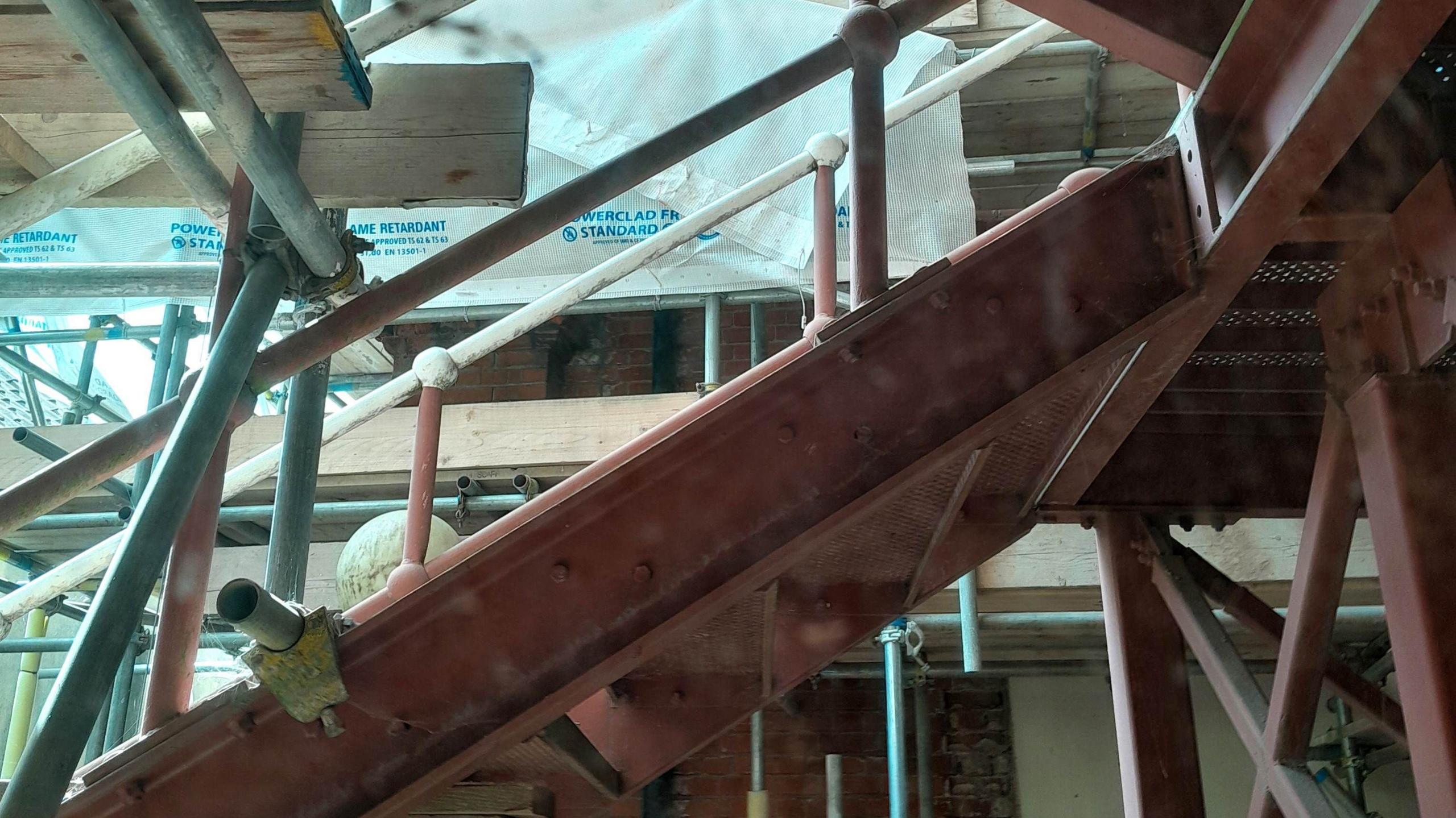 Close view of a dark wooded staircase leading up to another level. There are lots of metal poles, acting as scaffolding, around the room. 