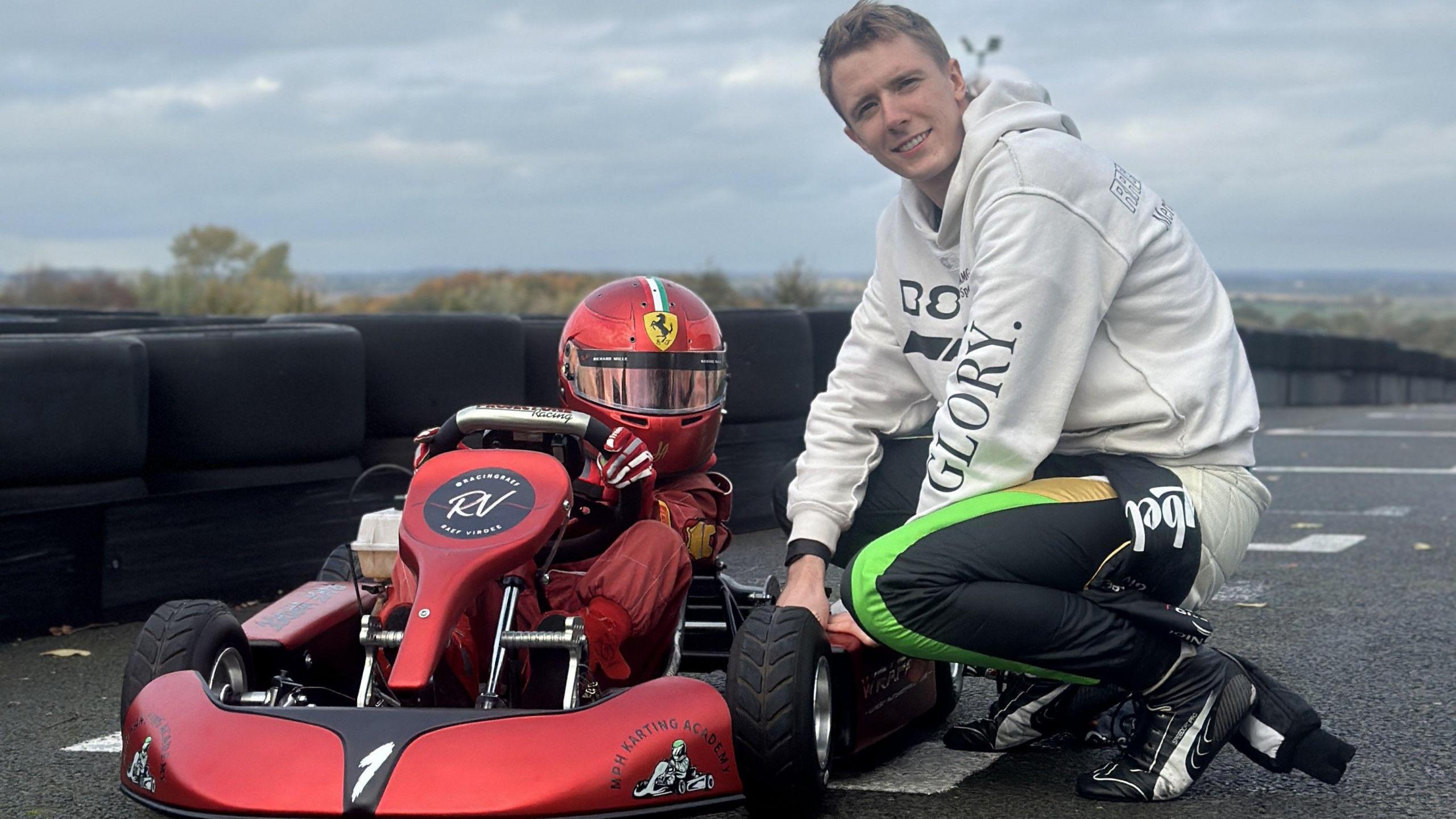 Raef is in his red racing suit and a red helmet, he is sitting on a red go kart. Kneeling next to him is Frederik Vesti, who is in a white hoodie and black trousers with a green stripe. He has fair hair.