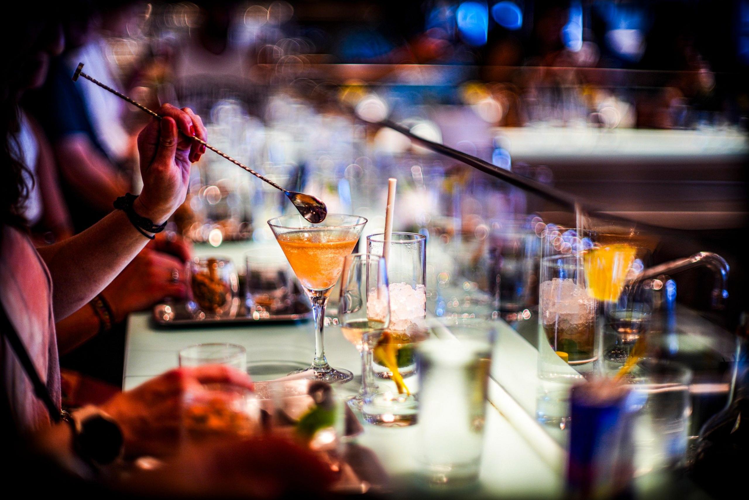 A person uses a long-handled spoon to mix a drink on a bar covered with drinking glasses. 