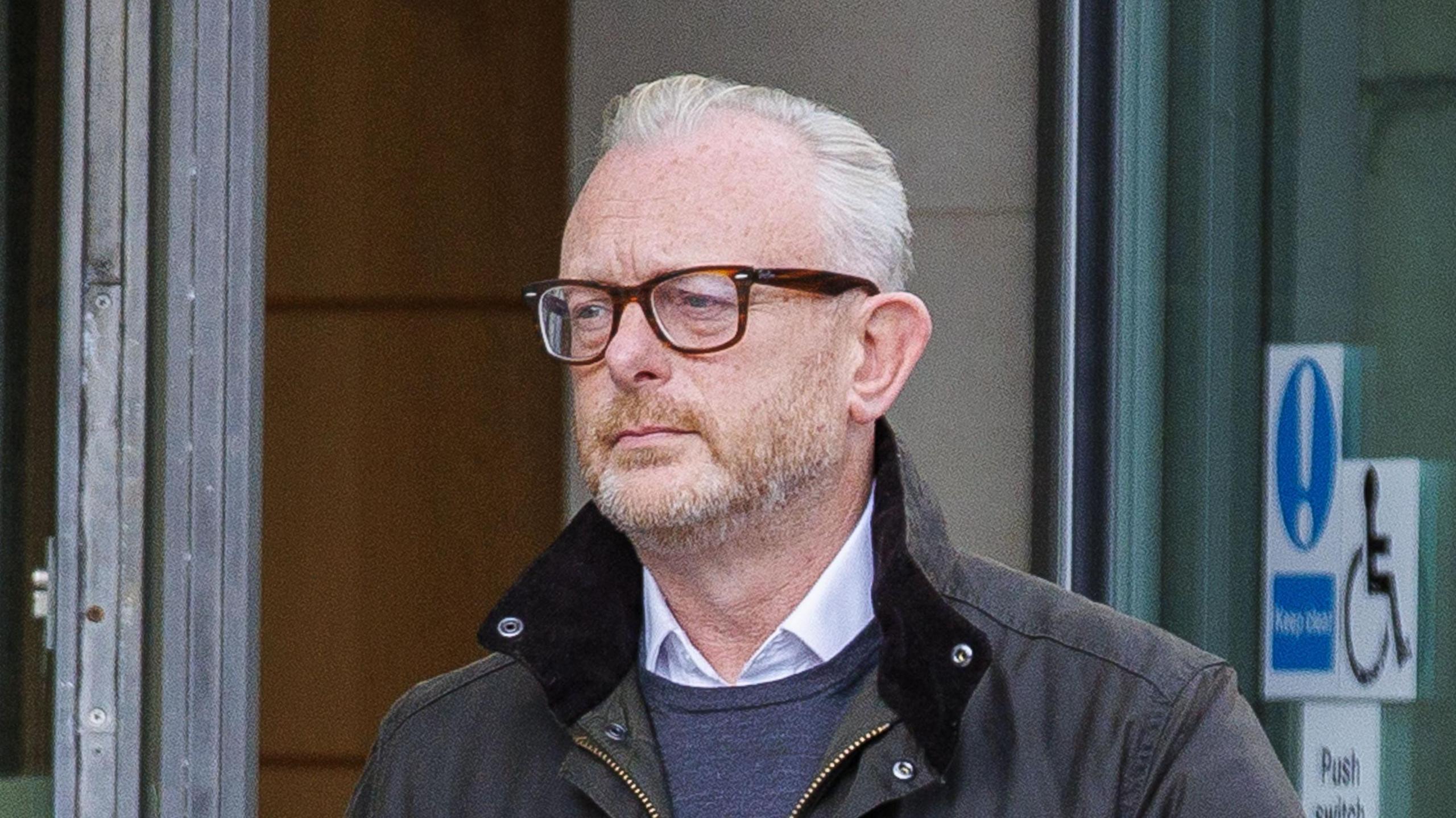 Ian Coulter is wearing a dark green coat with a upright brown collar. He has a grey jumper and a white shirt on. He is wearing brown glasses. He has shot facial hair and grey head hair. Behind him is a glass door with stickers on it.