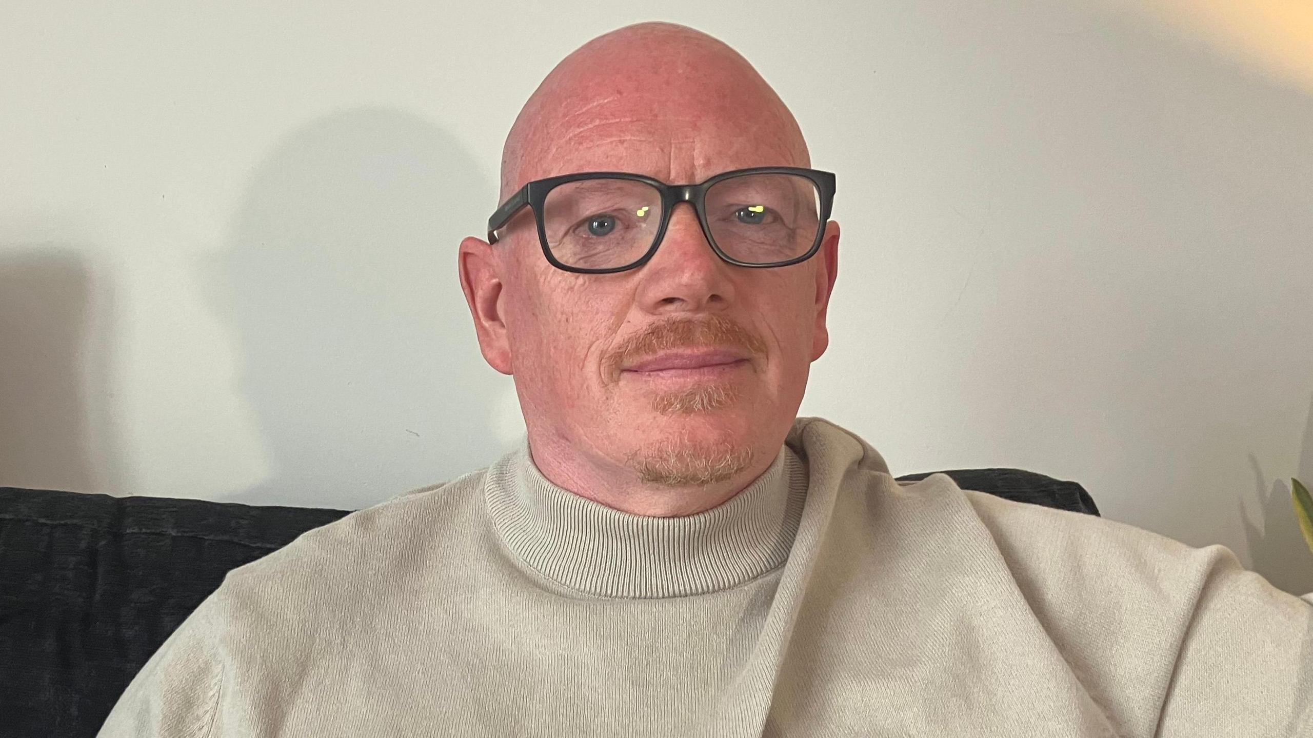 A man with glasses in a beige turtleneck sits on a sofa.