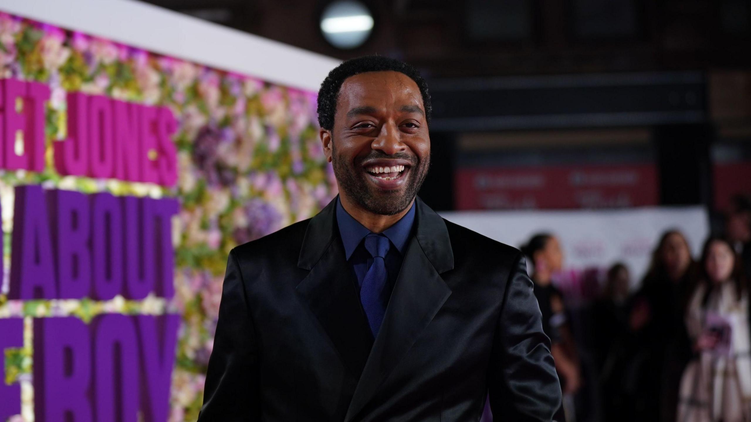 Chiwetel Ejiofor stands on a red carpet at a film premiere. He is laughing towards the camera and is wearing a black suit with a dark blue shirt and tie. A large wall covered in flowers is visible in the background along with other guests.