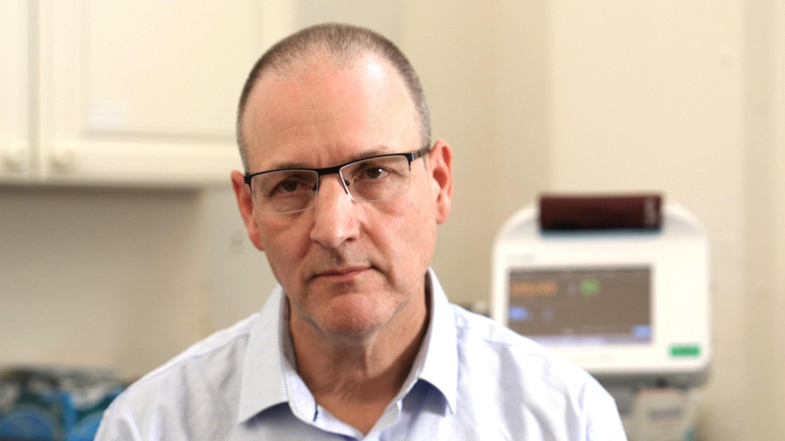 Martin Rolles looks at the camera. He wears a white shirt and glasses, and short dark hair. Medical equipment with a screen can be seen behind him.