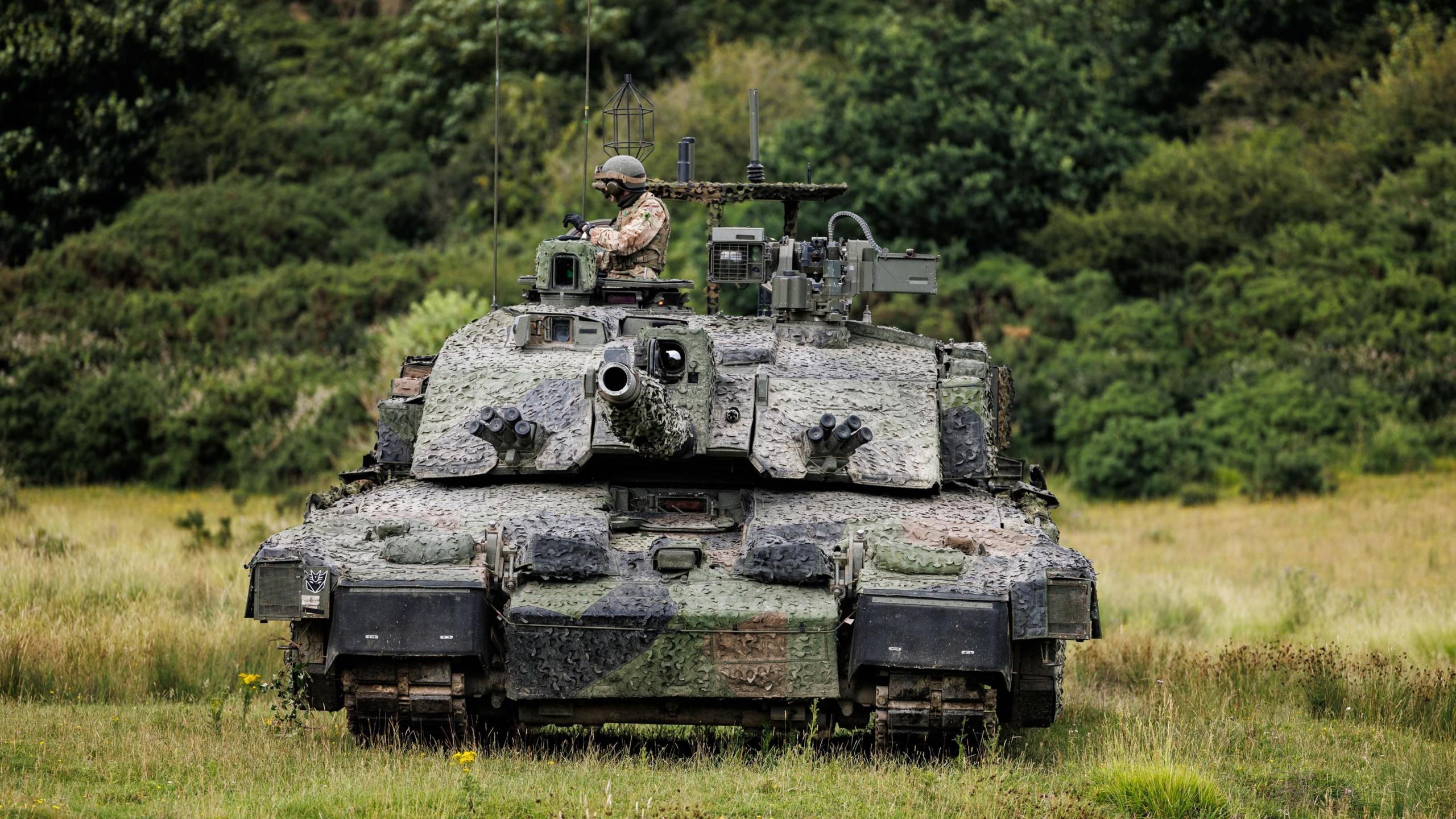 The Megatron variant of the Challenger 2 tank, heavily camouflaged, with a soldier wearing a helmet in the turret, advancing across a green field, with trees in the background