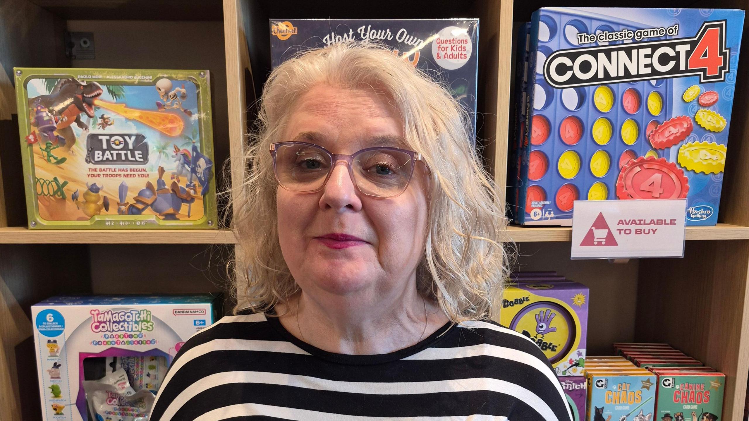 Kerry, a white haired woman in glasses, stands in front of shelf of toys in a shop, including Connect 4 and Toy Battle