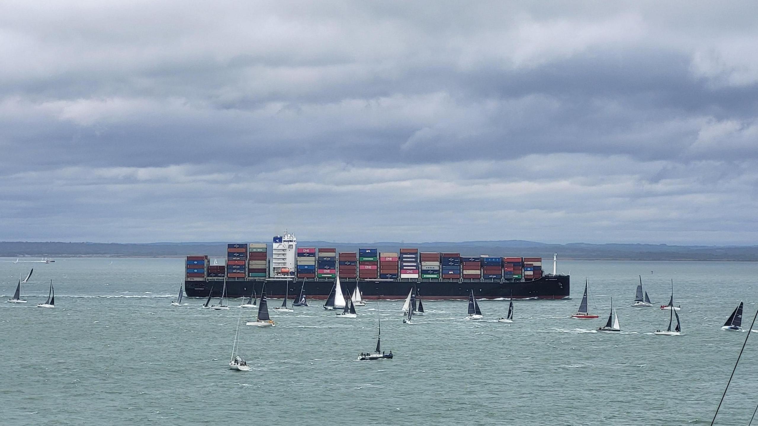 A large container ship loaded with crates. Smaller boats with sails are around it.