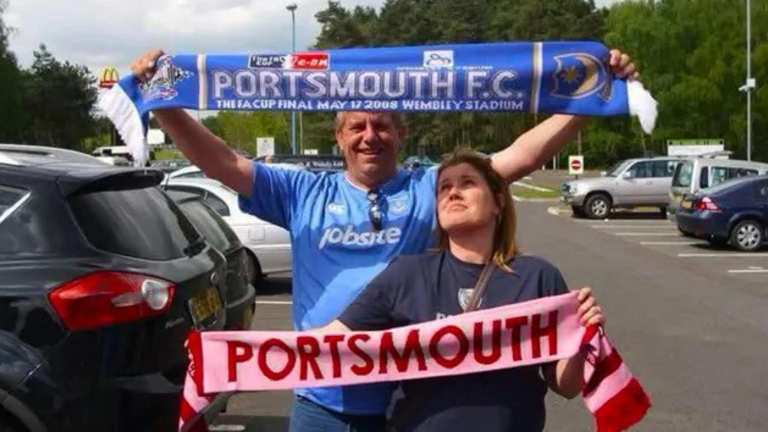 Alec Lumb stands in a car park with a woman. He is grinning widely at the camera, holding a Portsmouth FC FA Cup Final 2008 scarf above his head and wearing a pale blue Portsmouth FC t shirt and black sunglasses hooked in the collar. The woman is standing in front of him, she is looking up at his scarf and holds and pink and red scarf which reads 'Portsmouth'. She has brown hair, which is pushed away from her face with a pair of sunglasses and wears a dark blue short sleeved cotton t shirt.