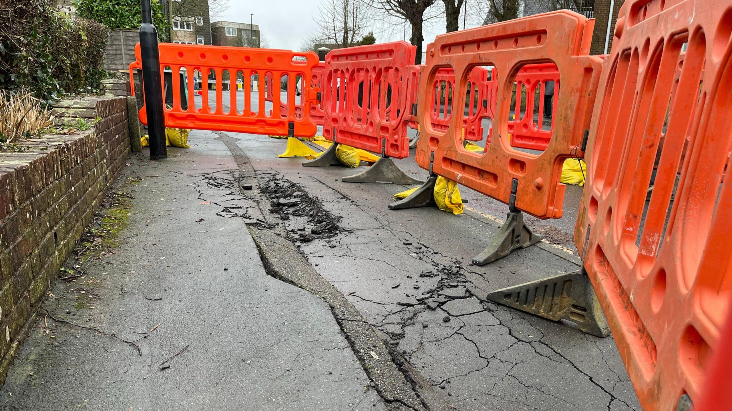 A pavement outside the surgery which is cracked and has become uneven. There is orange plastic fencing surrounding it with warning signs.