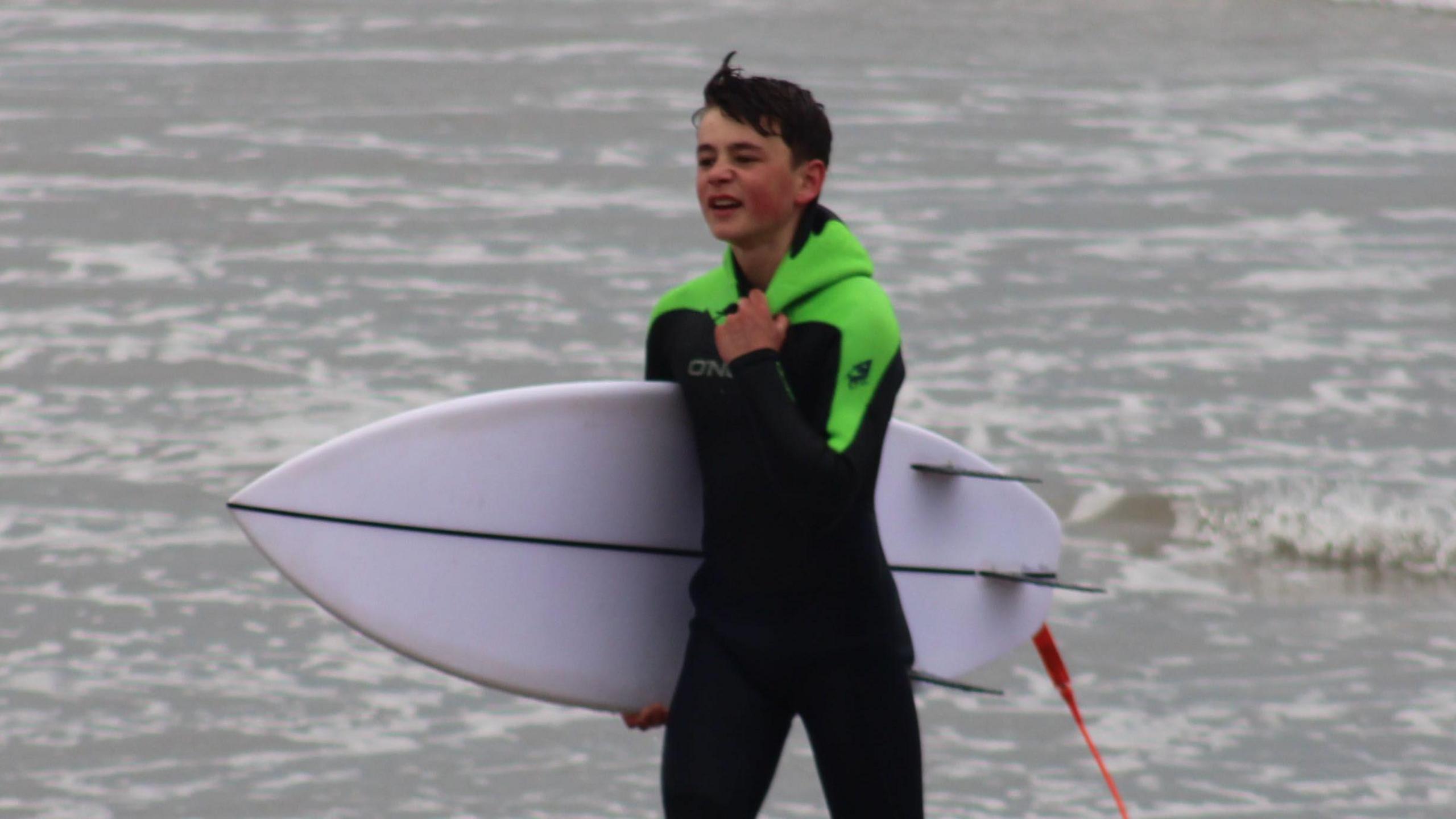 Oscar has reached the shore after the rescue and he has his short surfboard tucked under his right arm and his left hand is tugging on the neck of his wetsuit.