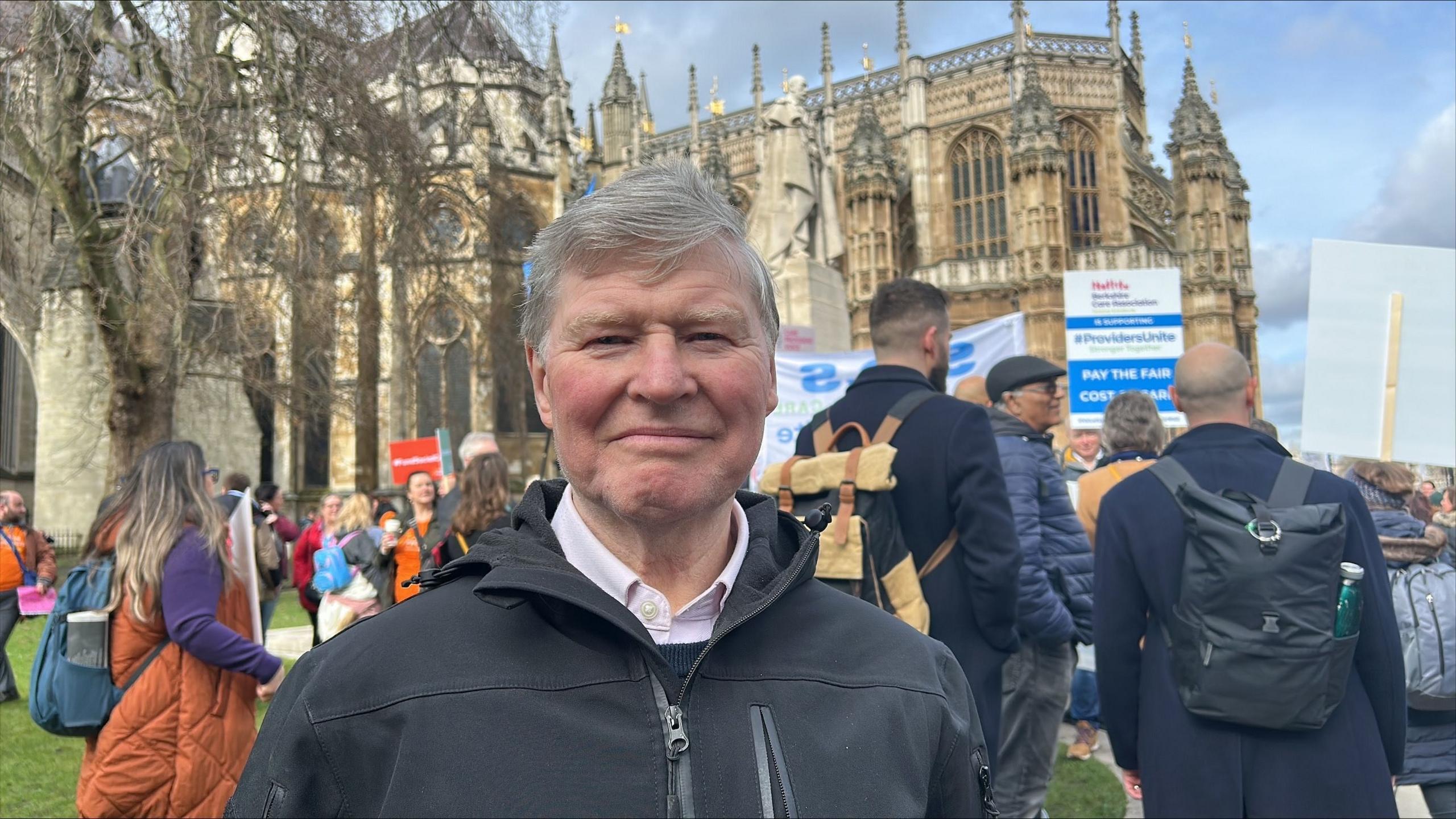 A grey-haired middle aged man with a pink shirt under a black jacket stands in front of protesters  holding placards at the Houses of Parliament in Westminster.