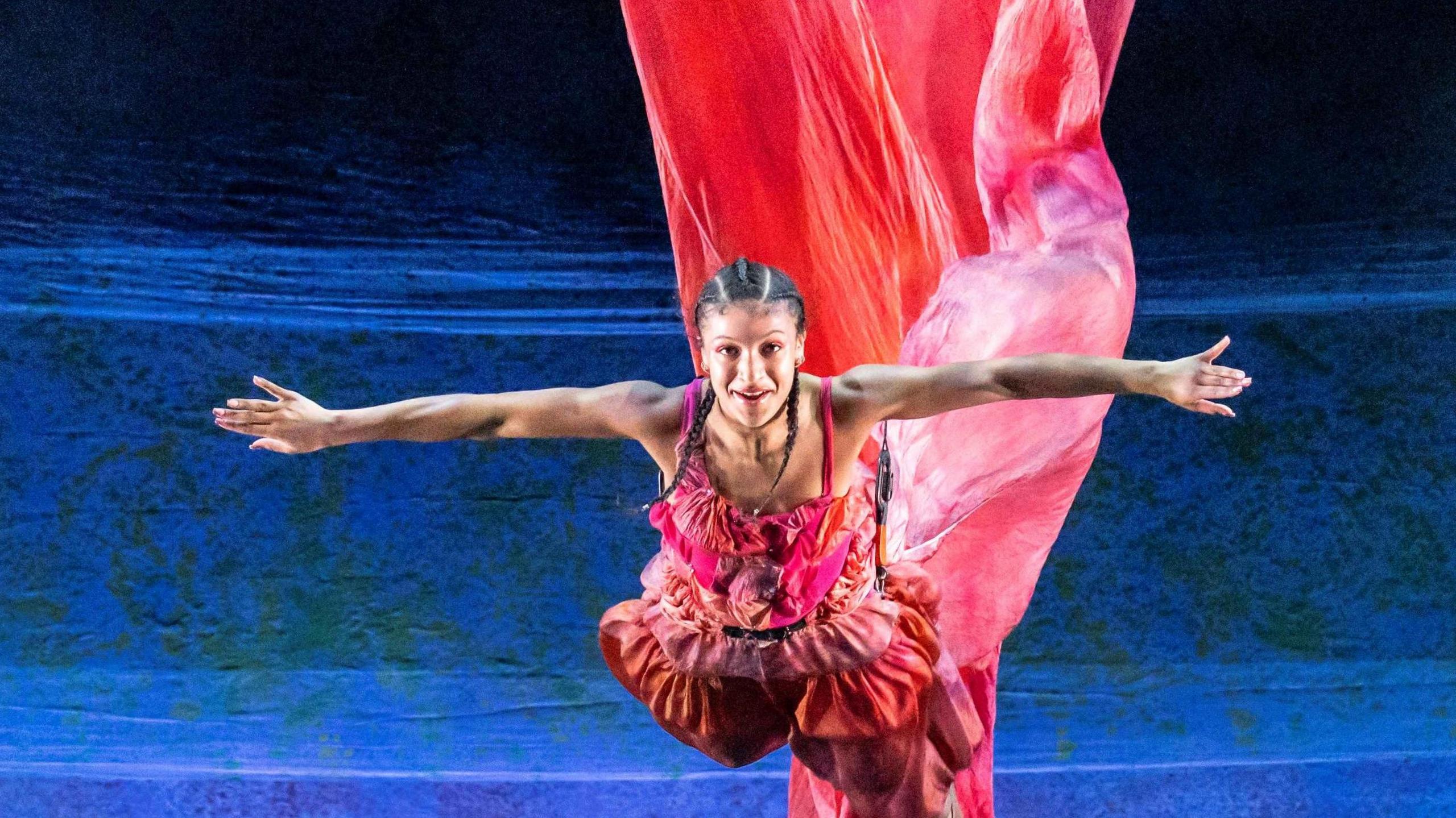 An actress starring in The Little Mermaid at the Bristol Old Vic is suspended from the roof. She is wearing a red and pink outfit, which is billowing up behind her against a deep blue background