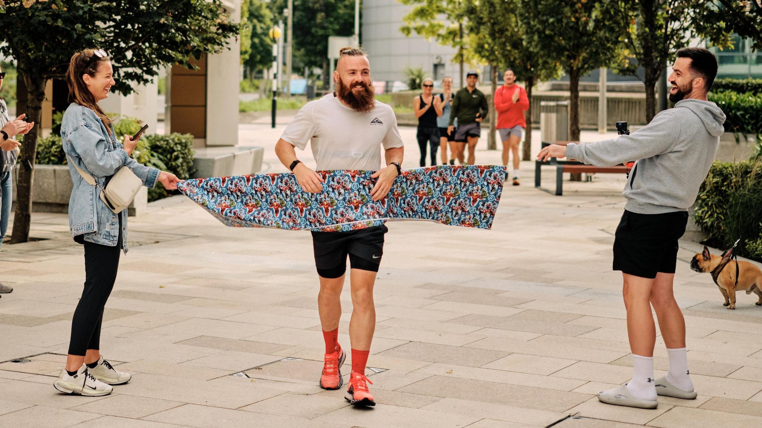 Joe smiles as he runs through the finish tape. He's wearing a white running top and people in the background are taking photos and clapping him. His hair is tied into a bun at the back of his head and his hair is shaved on the sides. He has a long beard as well.