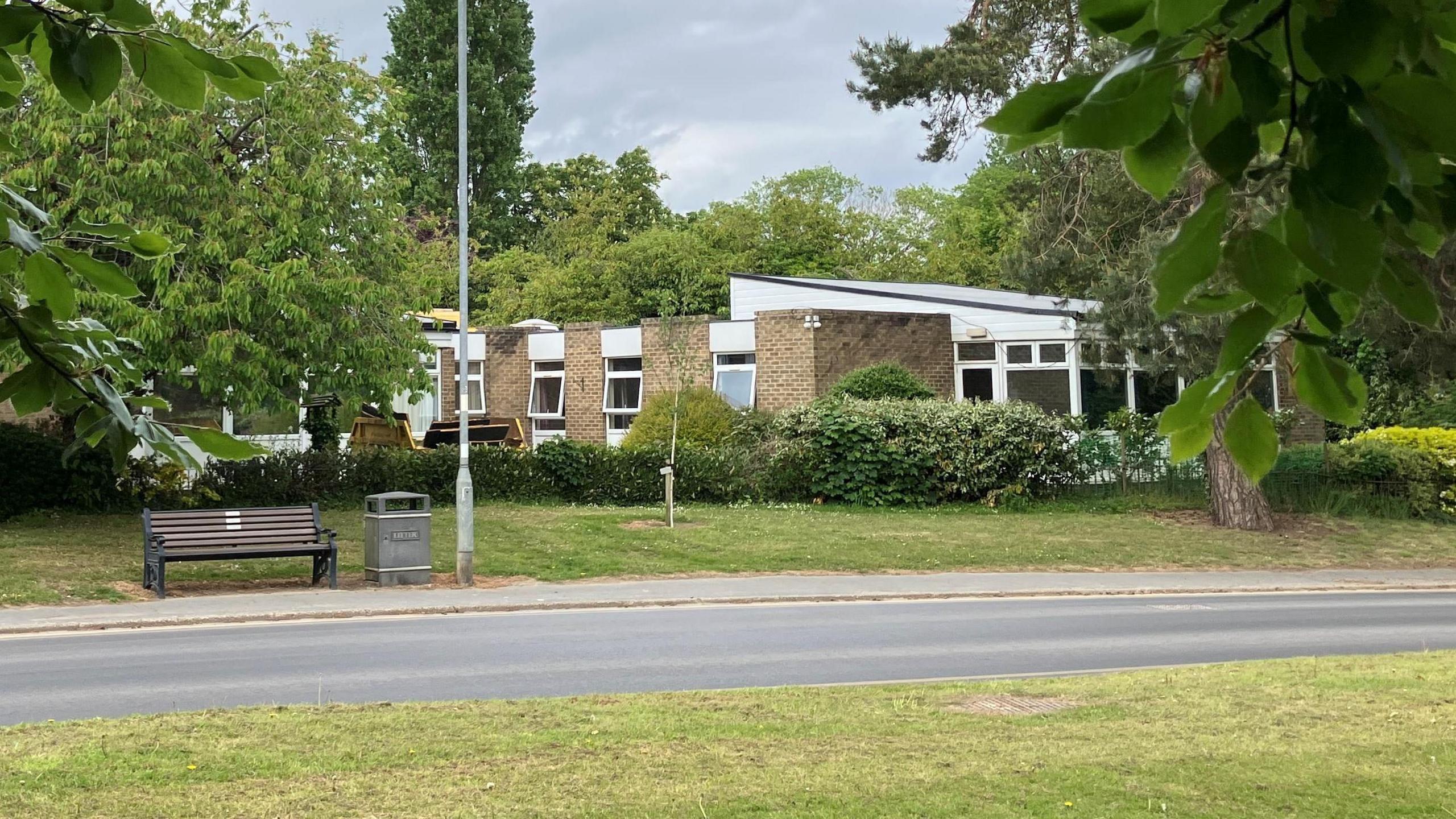 A brick one-storey building with windows and surrounded by trees. There is a road, a pavement, a bench, a waste bin and a lamp post in front.