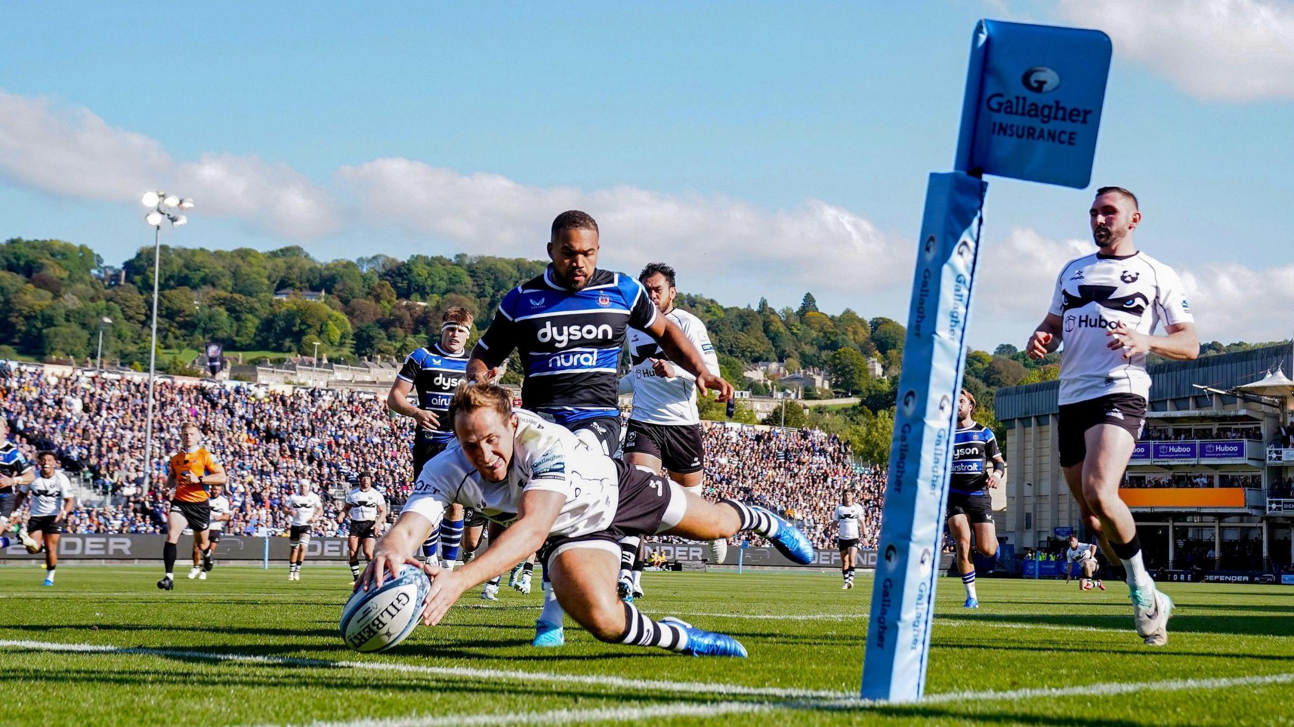 Bristol's Max Malins scores their first try during the Gallagher Premiership match at The Recreation Ground, Bath. He is diving over the line holding the ball while a packed stand is visible in the background, as are the hills of the city beyond