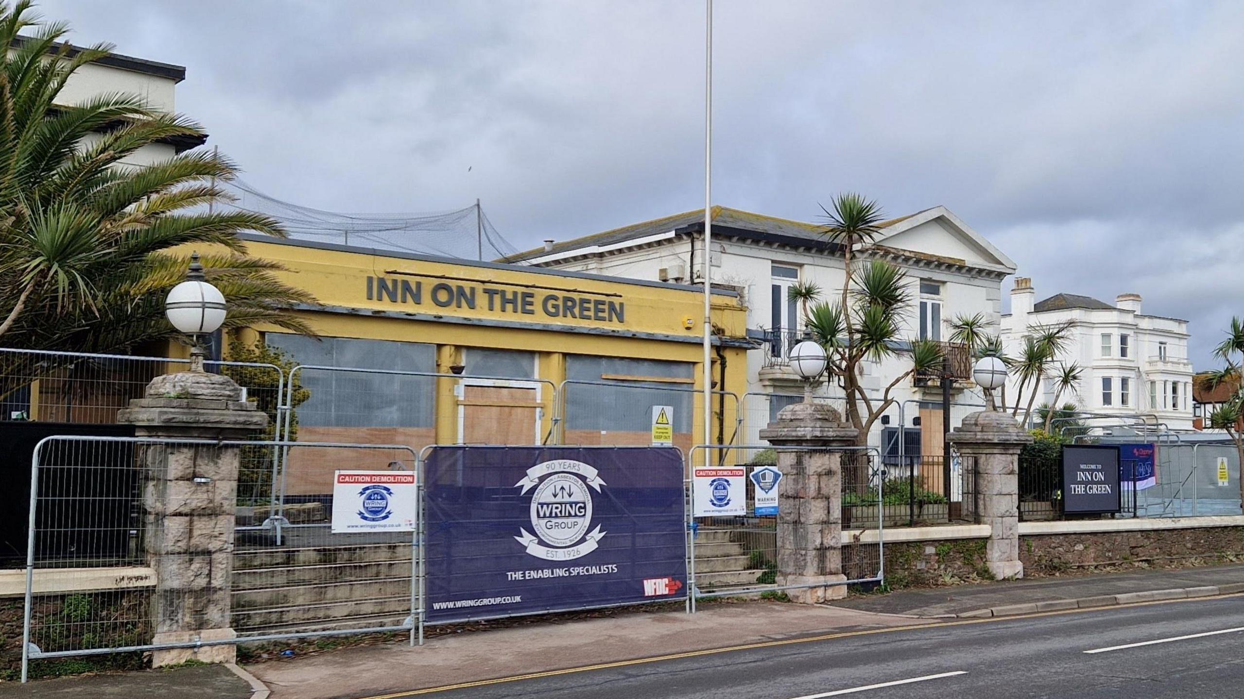 Railings around the Inn on the Green in Paignton. The pub is yellow and has its name written in large grey writing along the front. It is boarded up. There are palm trees either side of it and a large white building behind it. It faces onto a road.