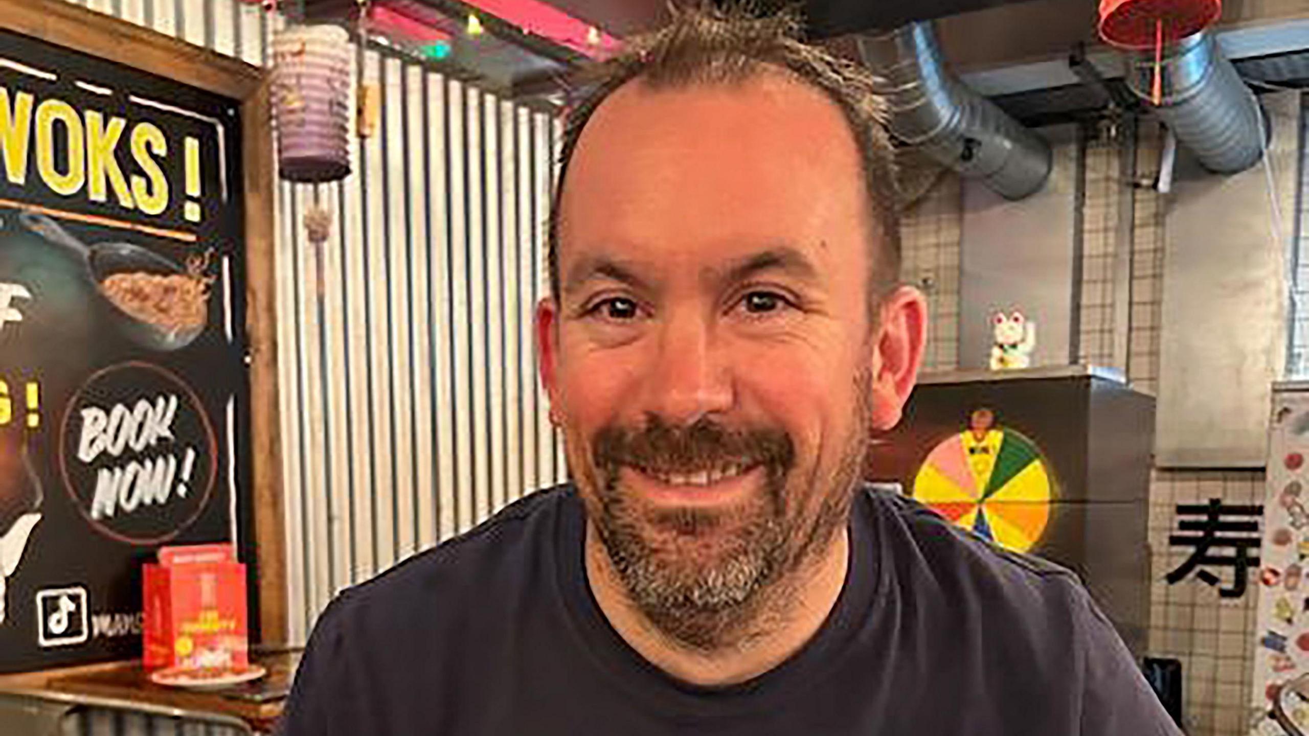 A man with short brown hair and a brown-grey beard sitting in a Chinese restaurant. He is smiling at the camera.