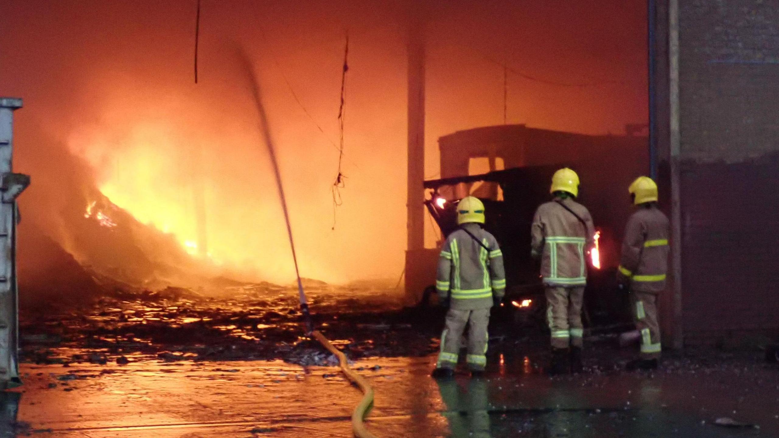 Three fire fighters are standing at the entrance to a warehouse with a large fire taking place inside it. A yellow hose is running along the ground and into the opening to the building, where it is spraying water towards the fire.