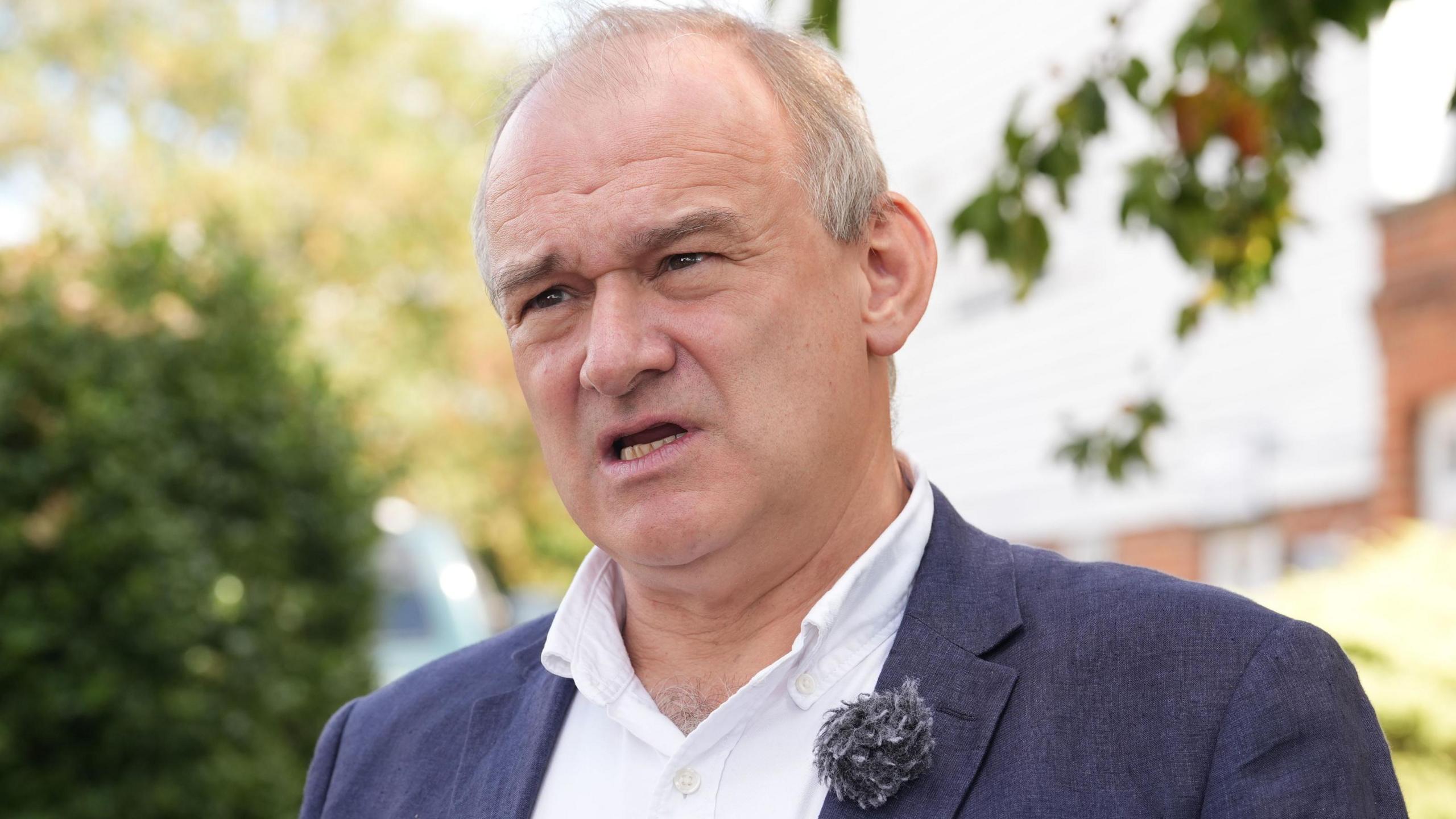Sir Ed Davey is wearing a dark blue linen suit and white open-necked shirt. He is standing outdoors in front of some trees. 