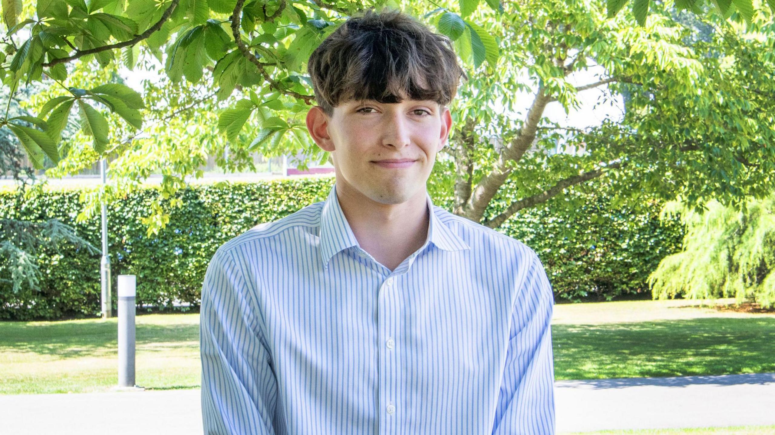 Felix Merritt wearing a blue and white pinstripe collared shirt and smiling at the camera. He has thick brown hair which covers his forehead, and is standing beside a concrete path below trees and low-hanging branches. It is a bright sunny day and the image looks warm.