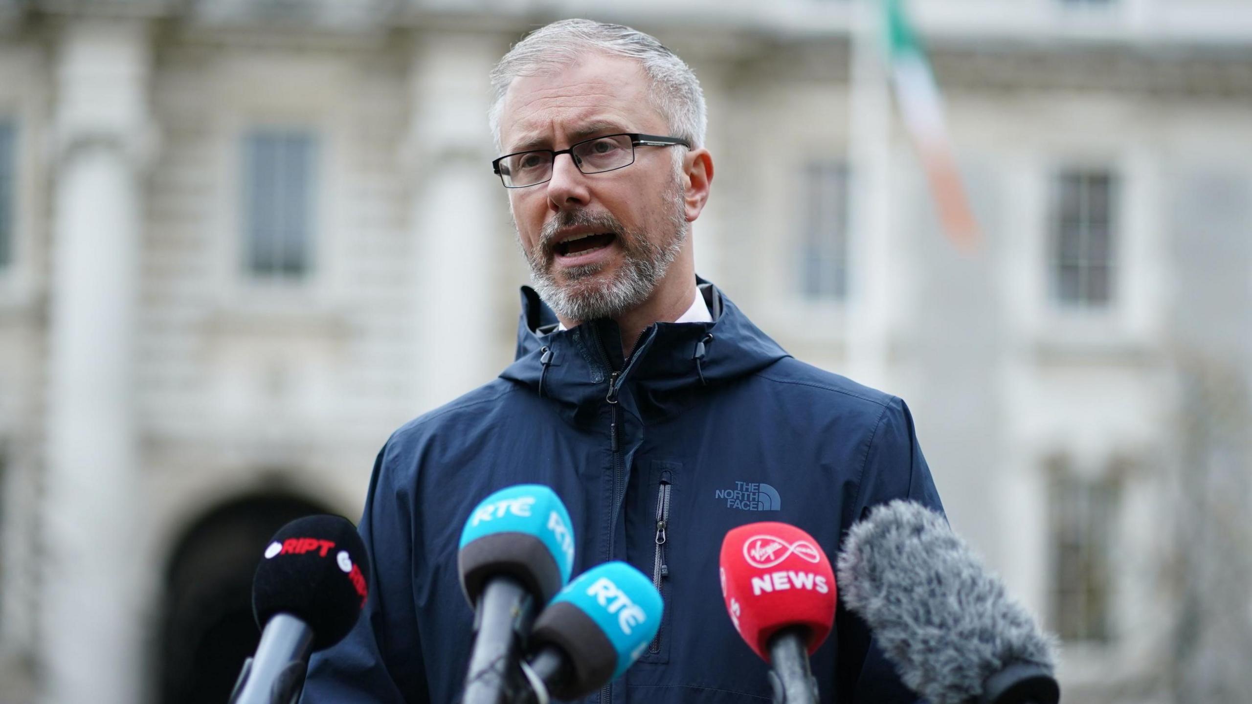 Man standing  and speaking at microphones. He is wearing glasses and a North Face jacket/ In the background there is a grey building and an Irish flag.