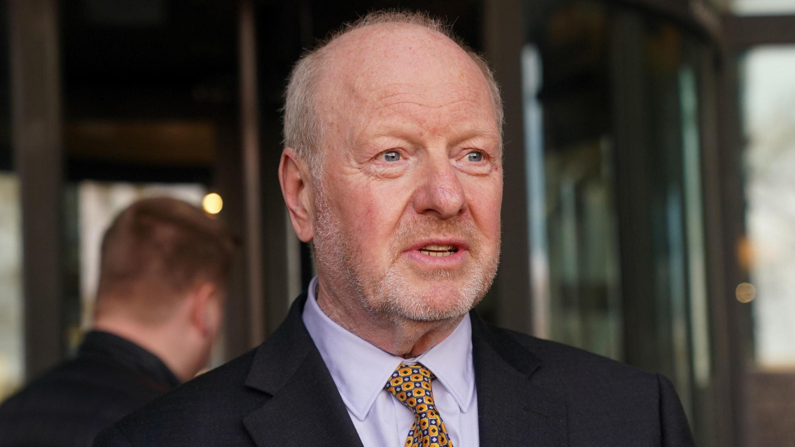 An older man wearing a suit with a lilac shirt and a blue and yellow tie