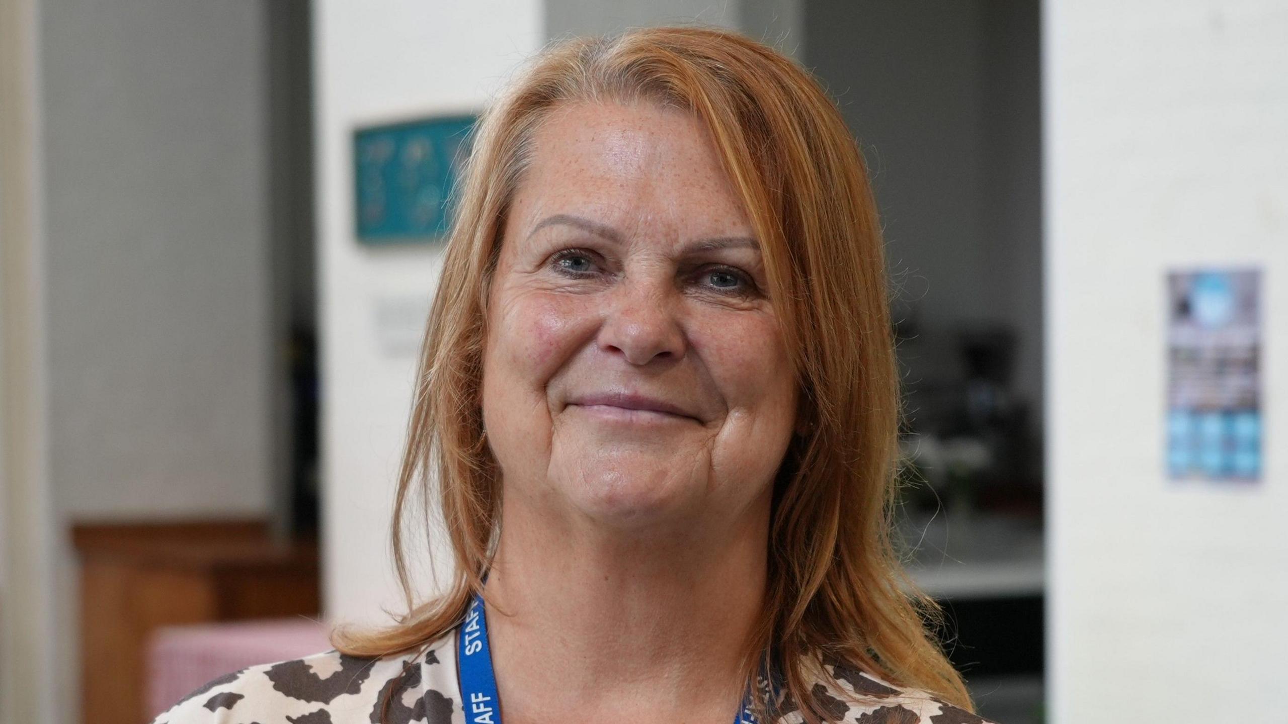 A woman with shoulder-length red hair smiles at the camera in a head and shoulders image. She is wearing a blue lanyard with STAFF written on the ribbon. 