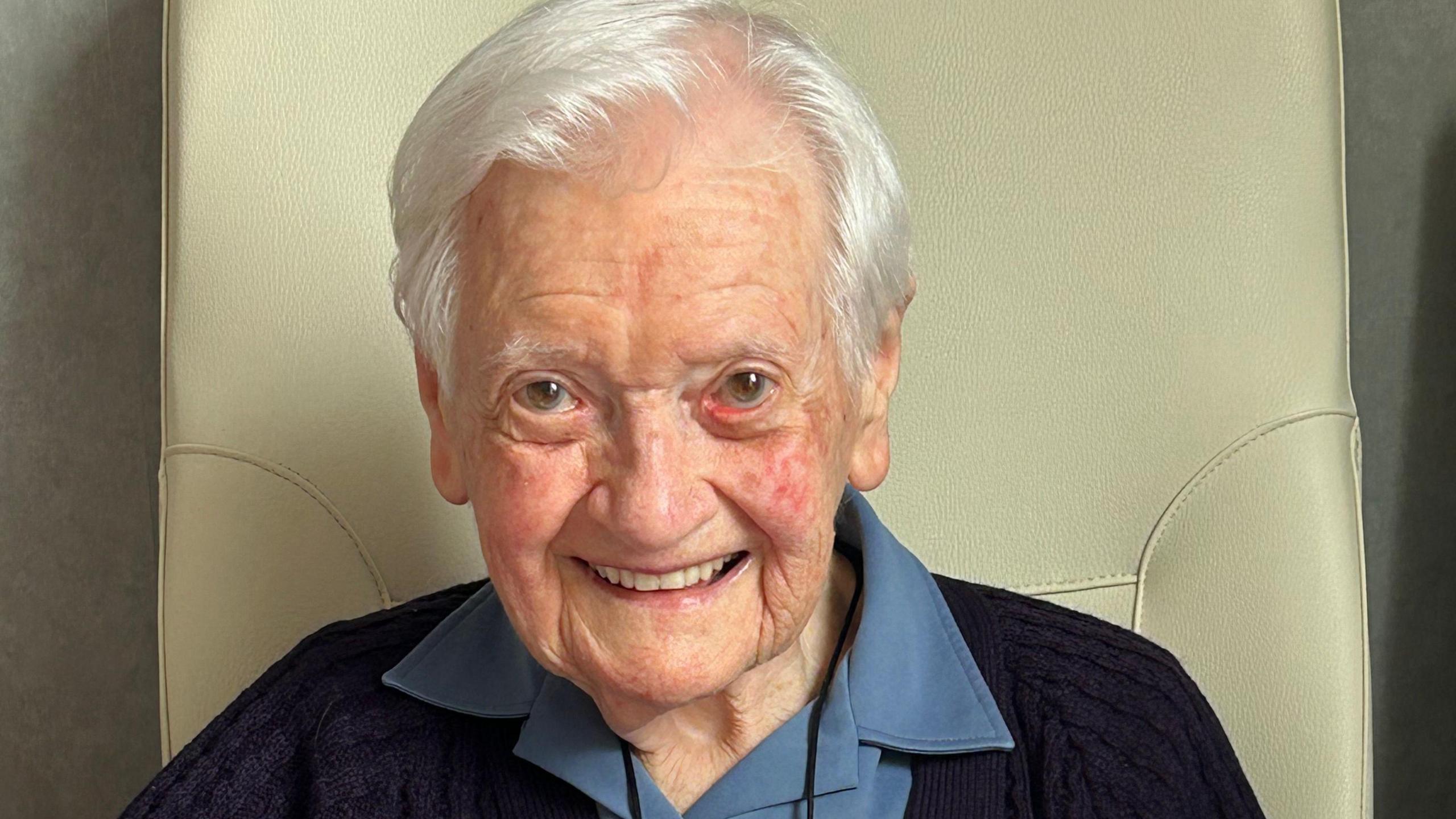 Sister Marie Louise sitting in a chair with a wide smile on her face. She has short white hair and brown eyes.