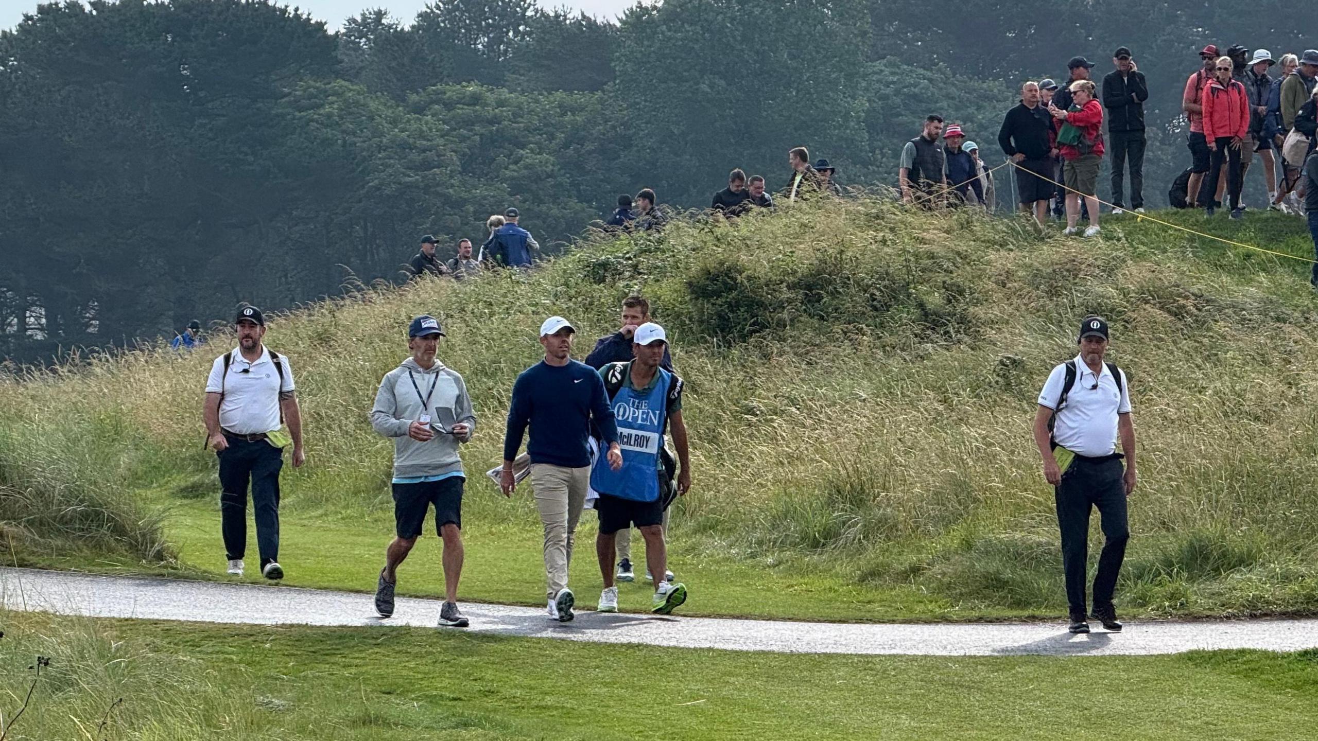 Men including a professional male golfer walk across a golf course carrying equipment. There are spectator standing up on a hill of grass watching them walk past.