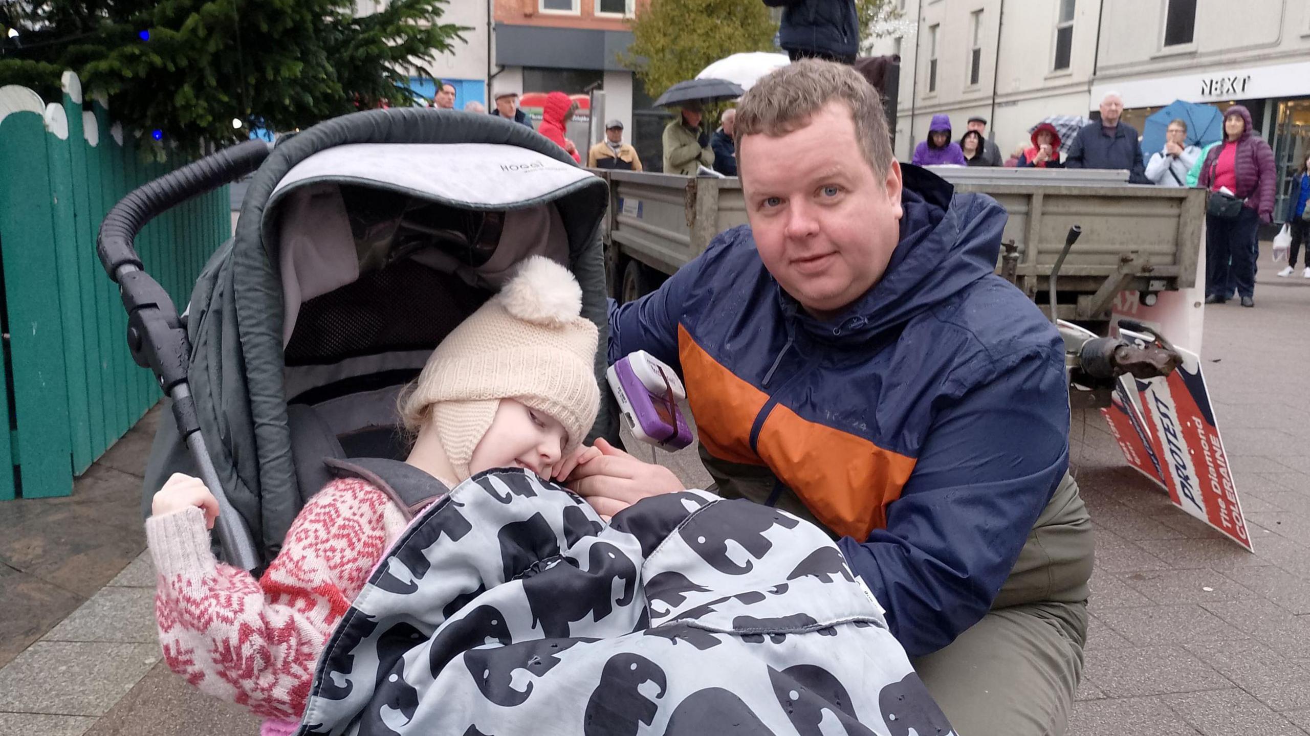 A child wears a beige woollen hat. She sits in a black buggy beside her father, who wears a navy and orange coat and green trousers. He has short brown hair. 