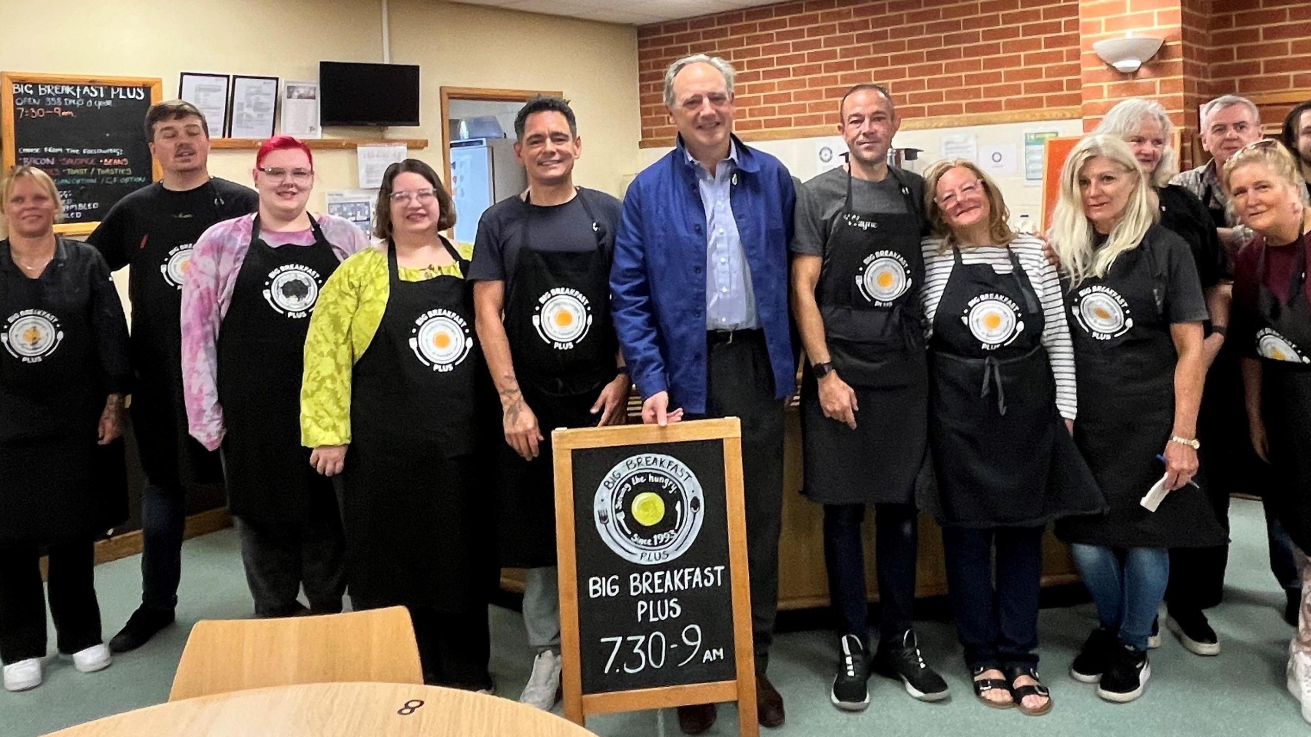 A group of people in a cafe-type area, all smiling at the camera. All but one man in the middle, the High Sherriff of Wiltshire, are wearing aprons with a logo that feature a large fried egg. There is a sign in between them saying Big Breakfast Plus.