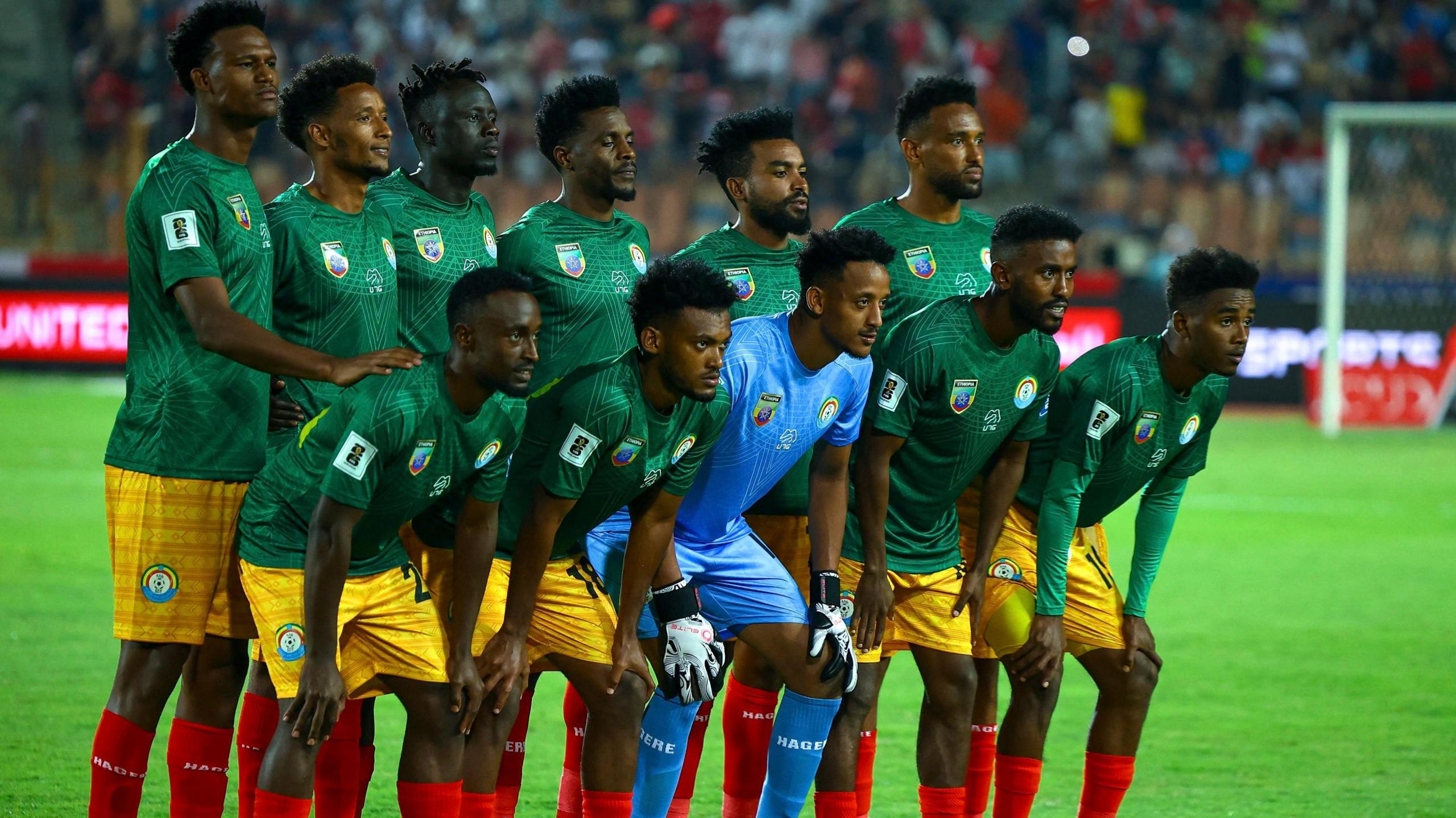 Ethiopia's football team, with outfield players wearing green shirts, yellow shorts and red socks and the goalkeeper in an all-blue kit, line up for a team photo before a match. A green pitch, goalposts and a packed stand can be seen out of focus in the background. Five players in the front row, with the goalkeeper in the middle, lean down and forward as they rest their hands on their knees, while the other players stand behind them
