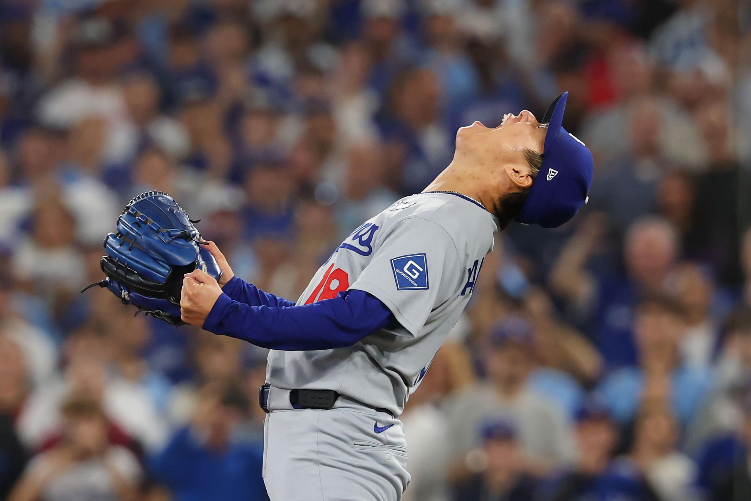 A baseball player wearing a gray uniform and blue cap stands on the field, leaning back with arms raised and glove in hand. The background shows a packed stadium with fans in blue and white, creating an energetic atmosphere.