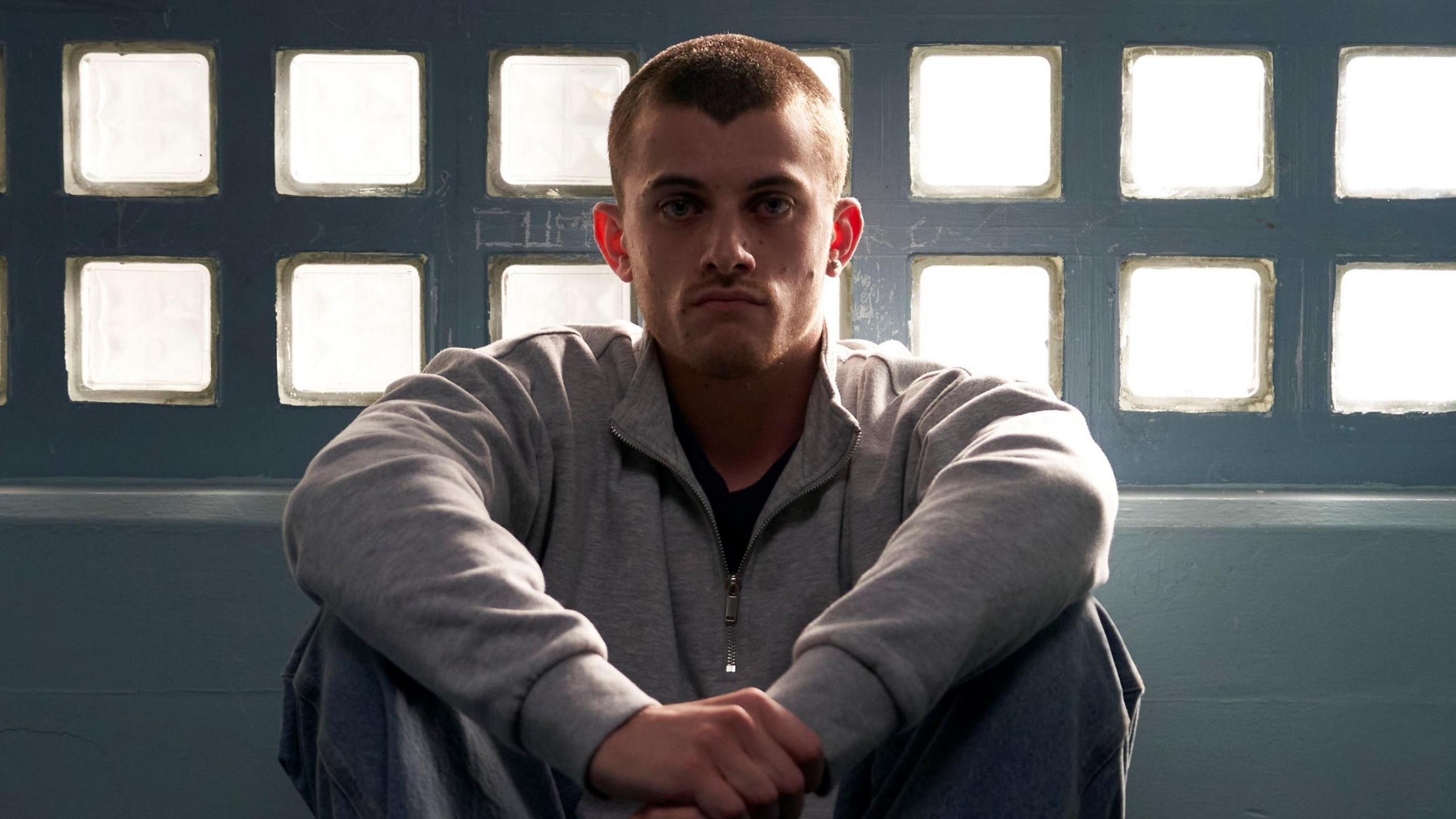 Jake Dunn plays Liam - he is pictured here sitting down, wearing a light grey top and dark trousers.