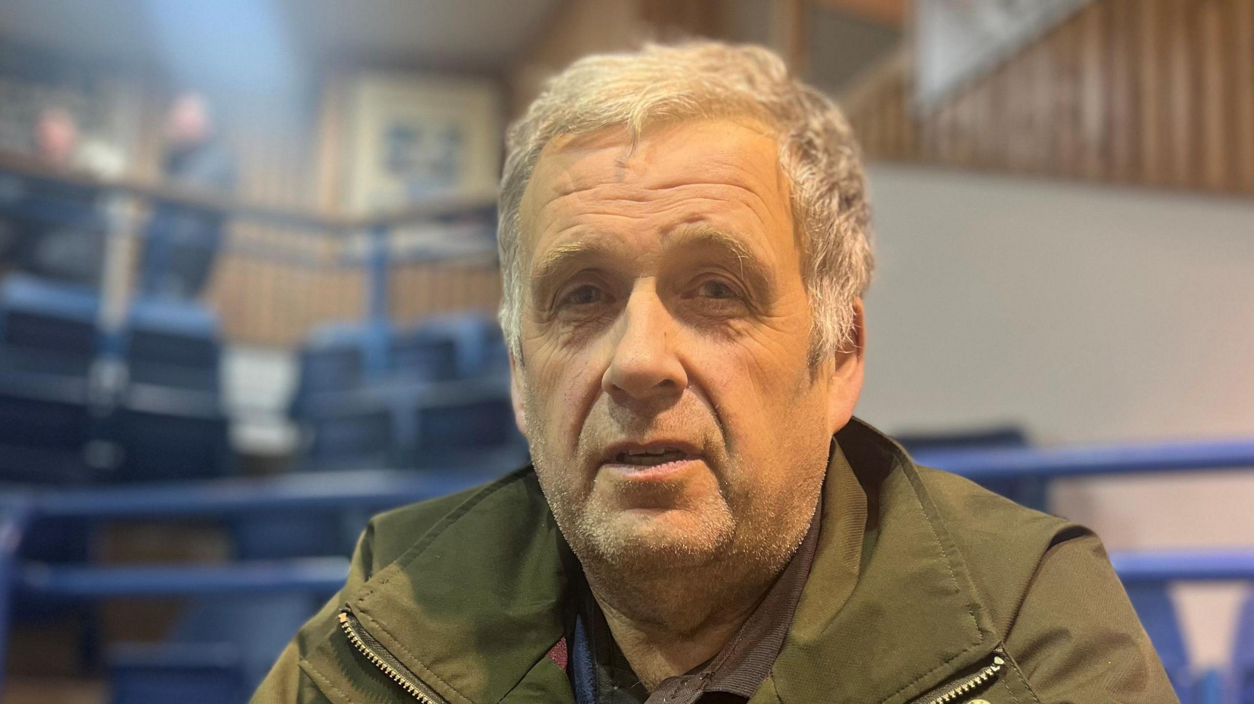 Man looking at the camera with short, grey hair. He is wearing a green wax jacket and is standing at an empty auction mart, featuring blue railings.