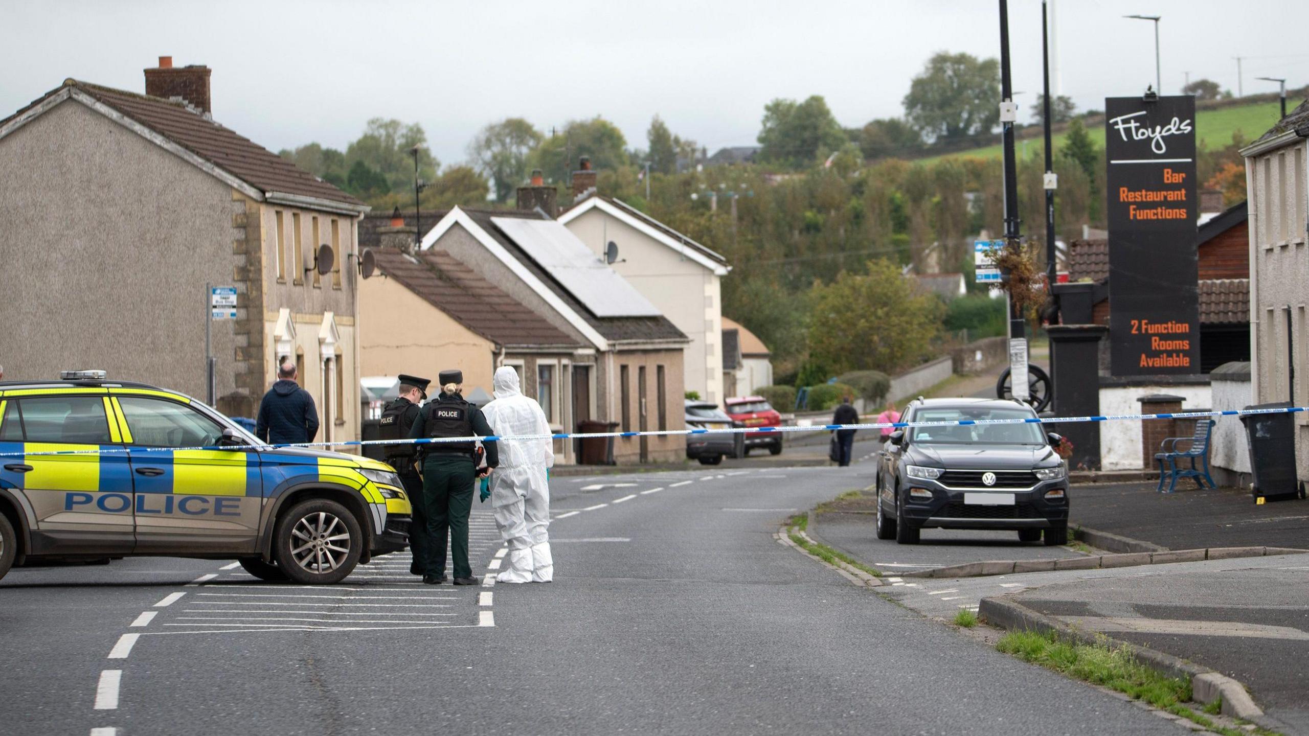 A number of police officers, one of which is wearing a forensic suit, stand behind a police tape. a police car is to their left, while a black car is to their right. A number of shops and buildings can be seen in the background, as can more vehicles, and another line of police tape where another police officer is standing.