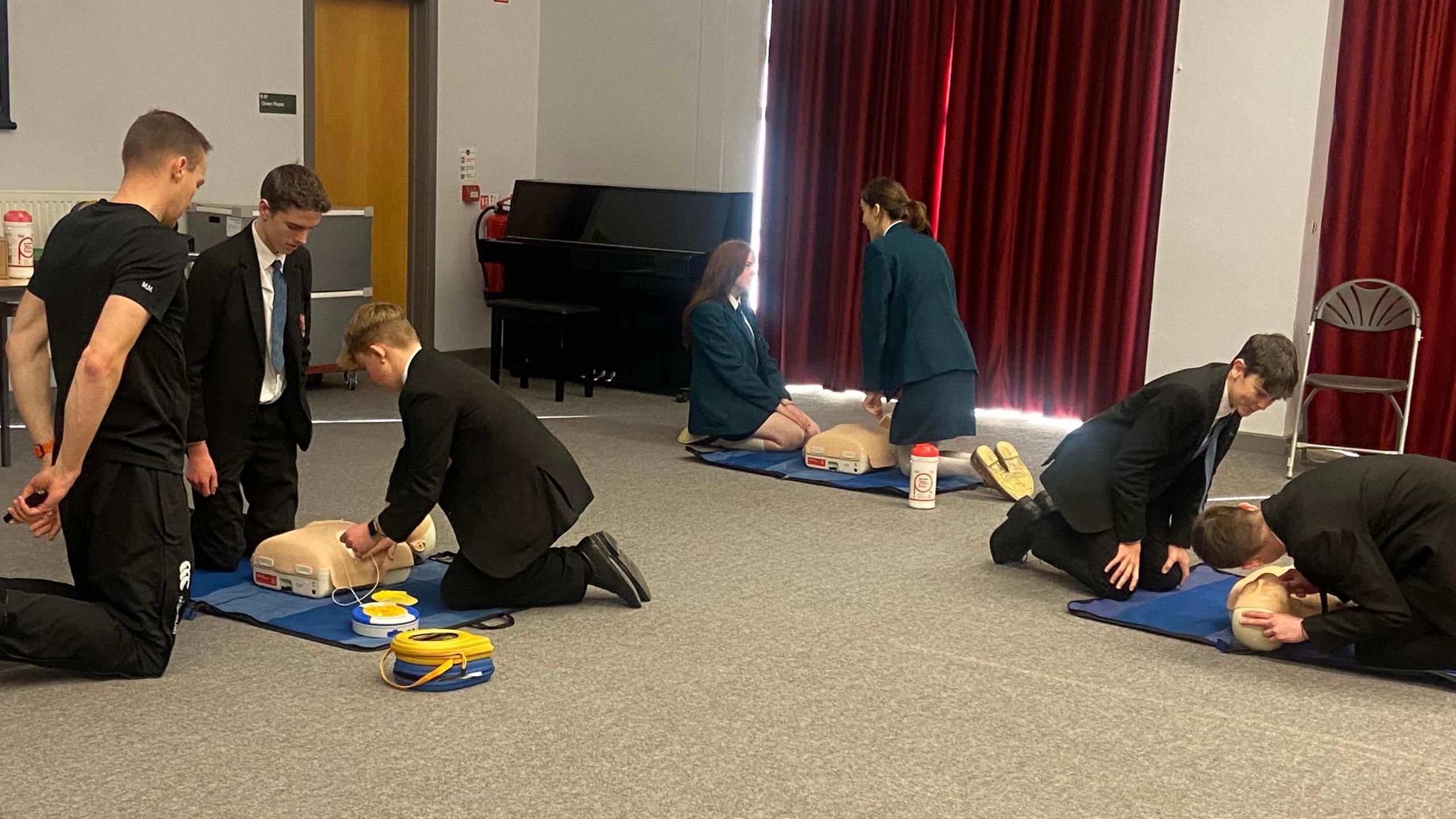 Groups of school students performing CPR on practice dummies. Teacher with a group of two boys to the left, group of two girls in the middle, and group of two boys to the right. The group to the left are using a practice defibrillator.