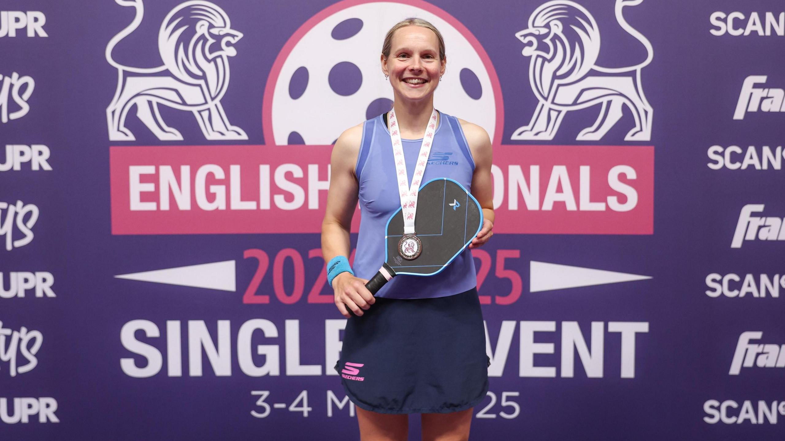Thaddea is stood smiling at the camera, holding her racket across her body while a medal hangs from her neck.