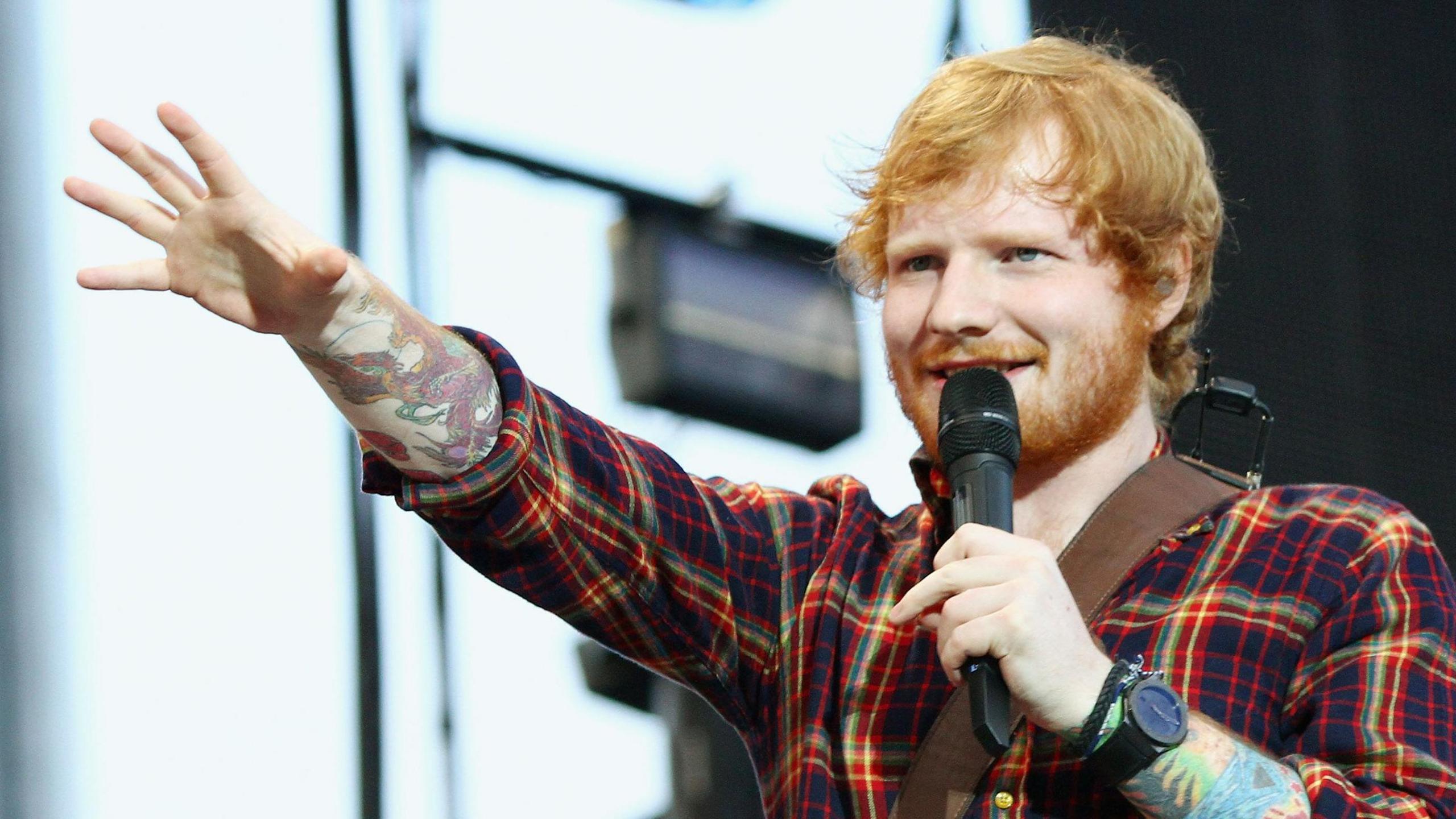 A ginger haired singer wearing a red checked shirt singing into a mic with tattoos up his arm.