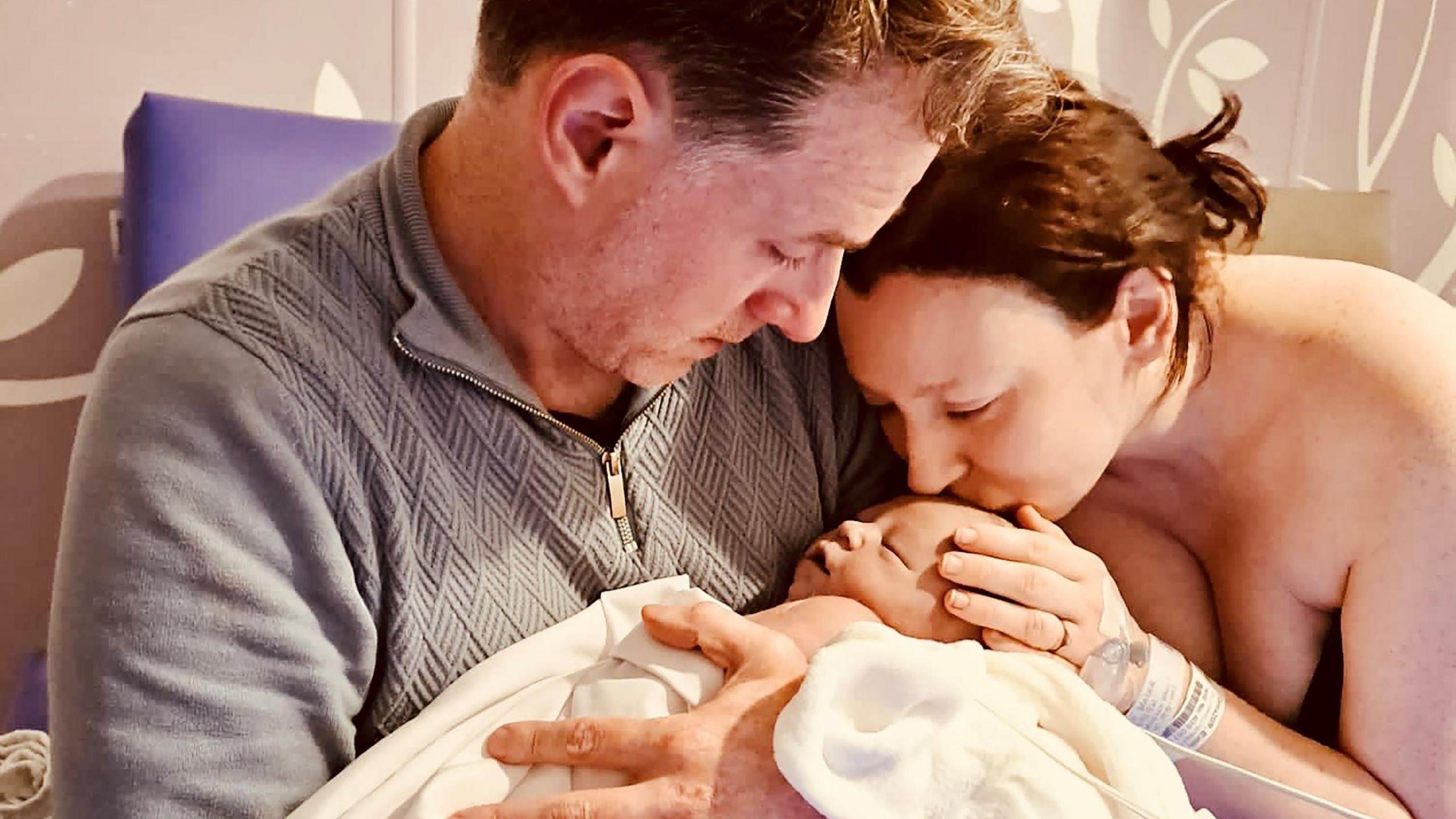 Rhodri Thomas holds baby Liliwen in his arms while his wife Emily Brazier kisses her forehead with one hand on her head.