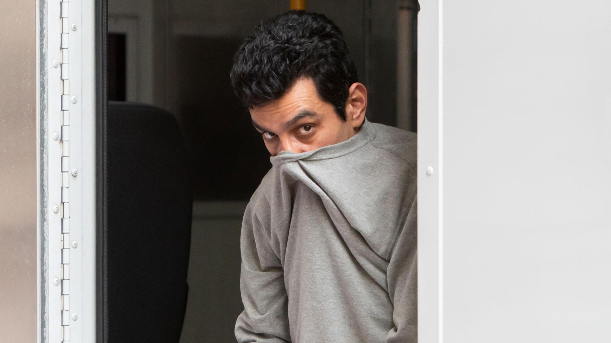 Alireza Askari leaving a police van. He wears a grey sweatshirt which is pulled above his nose to cover his mouth. His head is lowered and he has dark black hair, brown eyes and his eyes look up at the camera.