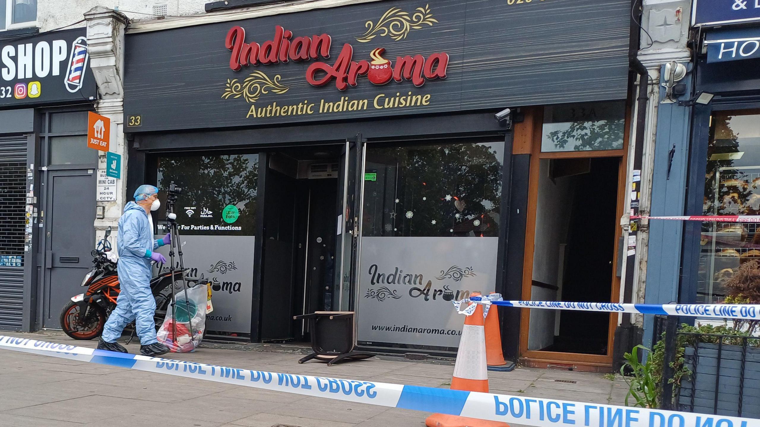 A forensic investigator outside a restaurant in Ilford. In front of the restaurant is police tape.