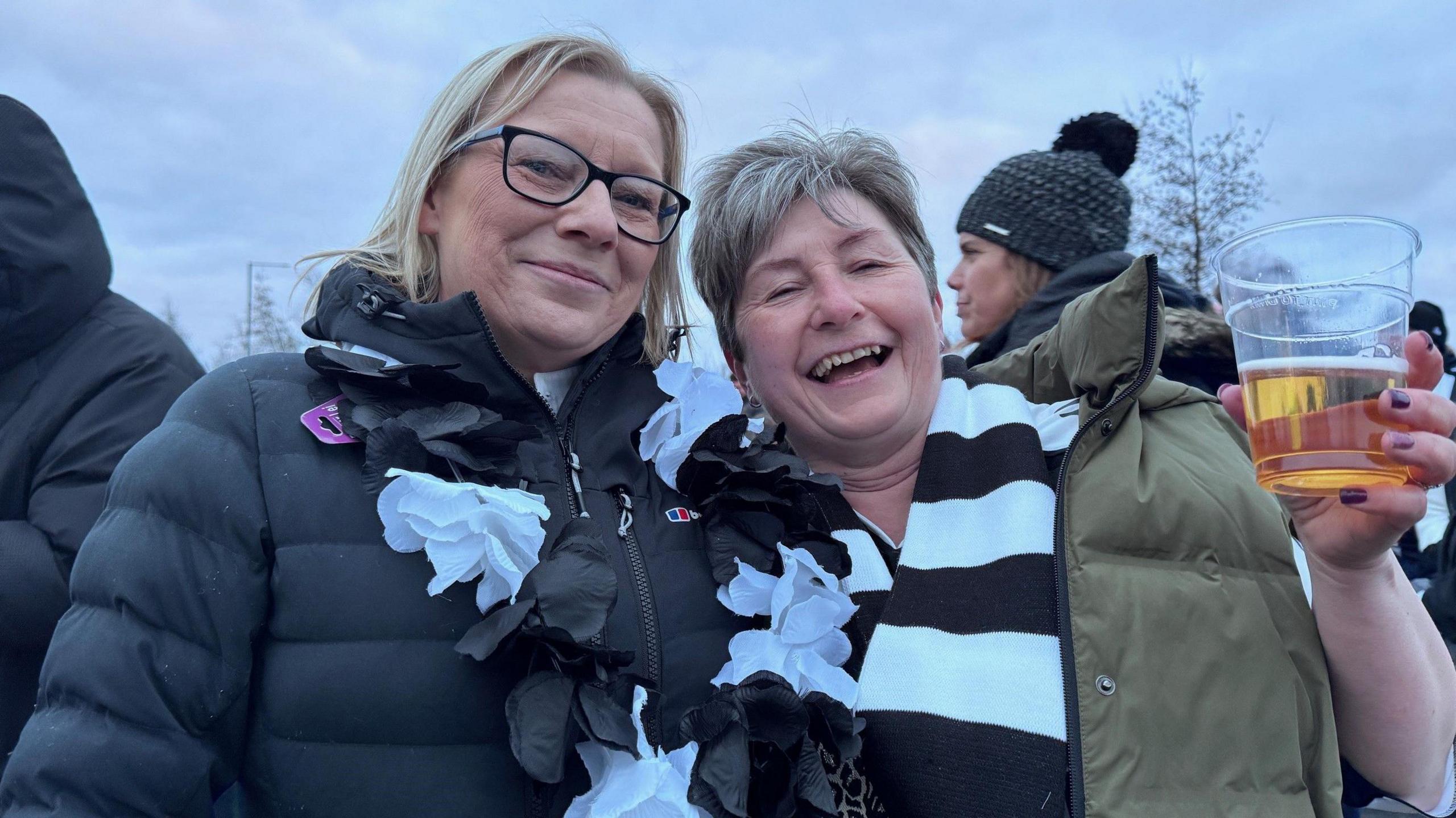 Two women celebrating at the Newcastle fan zone. Both are wearing black and white scarves. The one on the right is holding a glass of beer.