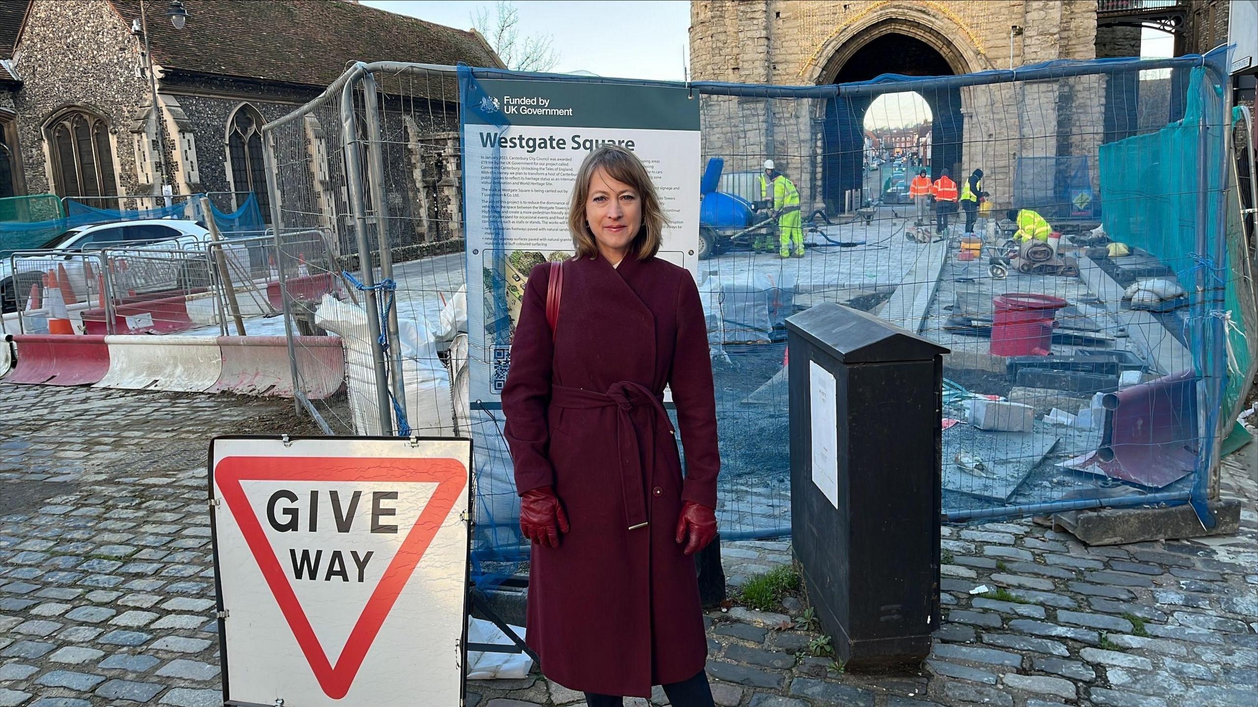 A woman wearing a dark coat and gloves standing in front of roadworks in front of Westgate Towers in Canterbury with a metal fence and workmen behind her